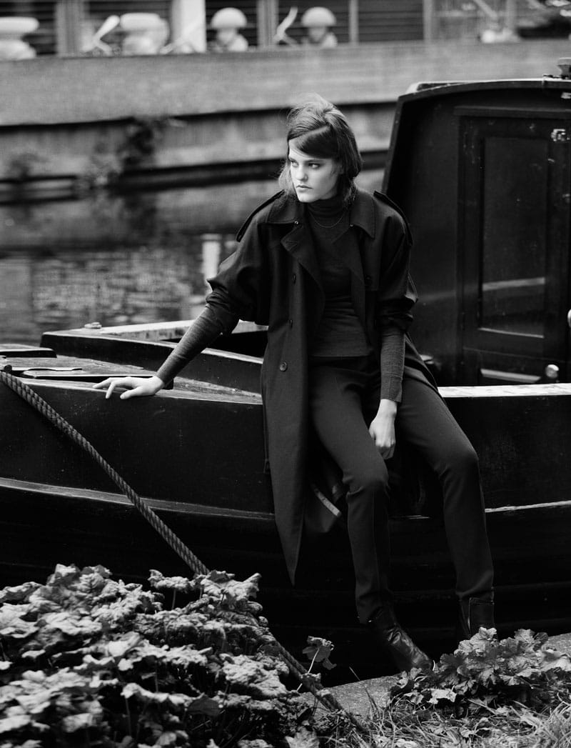 Kia Low seated on a black canal boat in a Paul Smith trench coat and boots with Viktor & Rolf trousers.