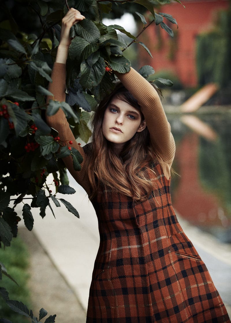 Kia Low reaching into leafy branches in a rust-and-black tartan Bimba & Lola look.
