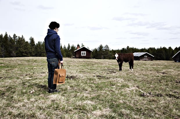 Sandqvist Fall/Winter 2011: Person with tan leather tote facing a cow near rural cabins.