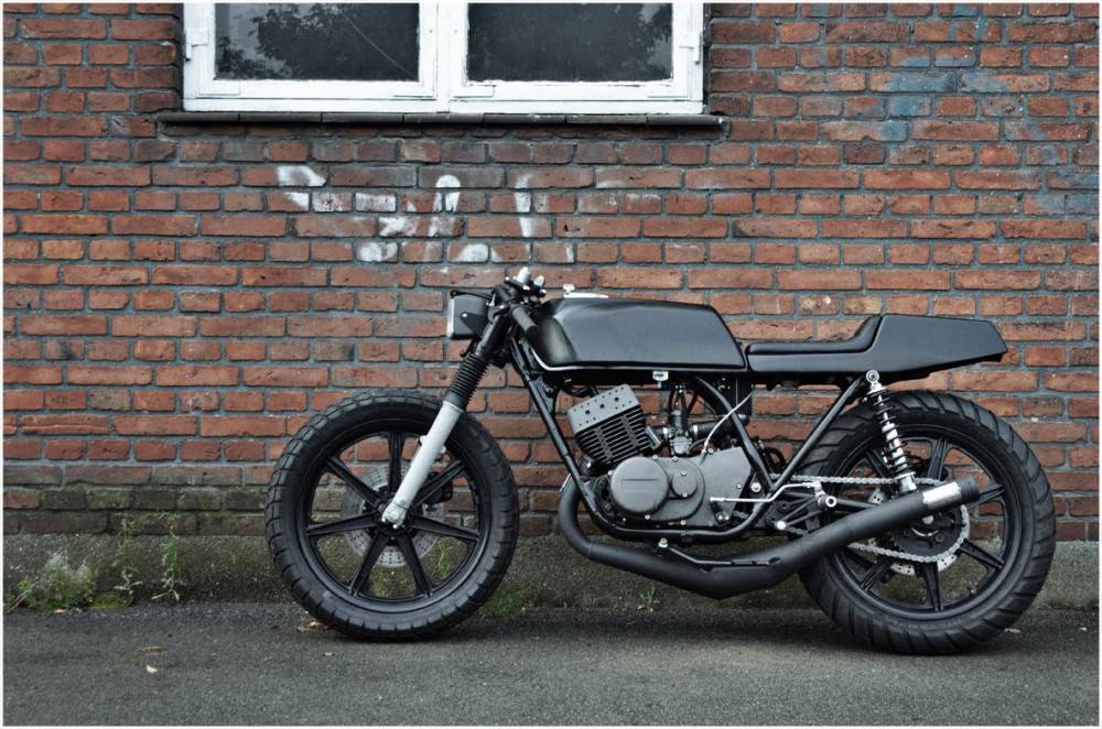Left-side profile of the black Wrenchmonkees Yamaha RD400 café racer against a brick wall.