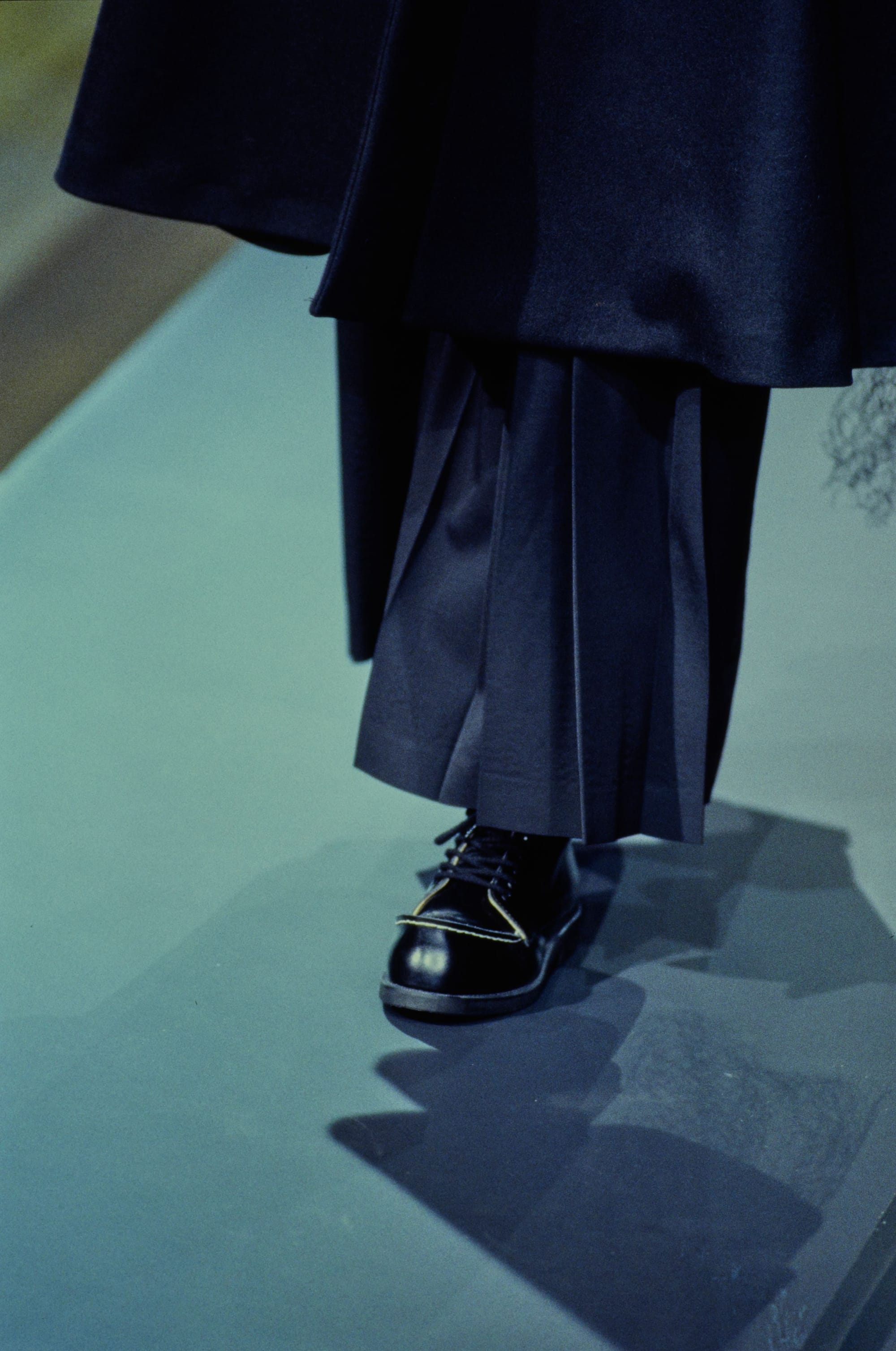 Yohji Yamamoto Fall 1995 detail 3: Pleated navy garment hem above a glossy black lace-up shoe on the runway.