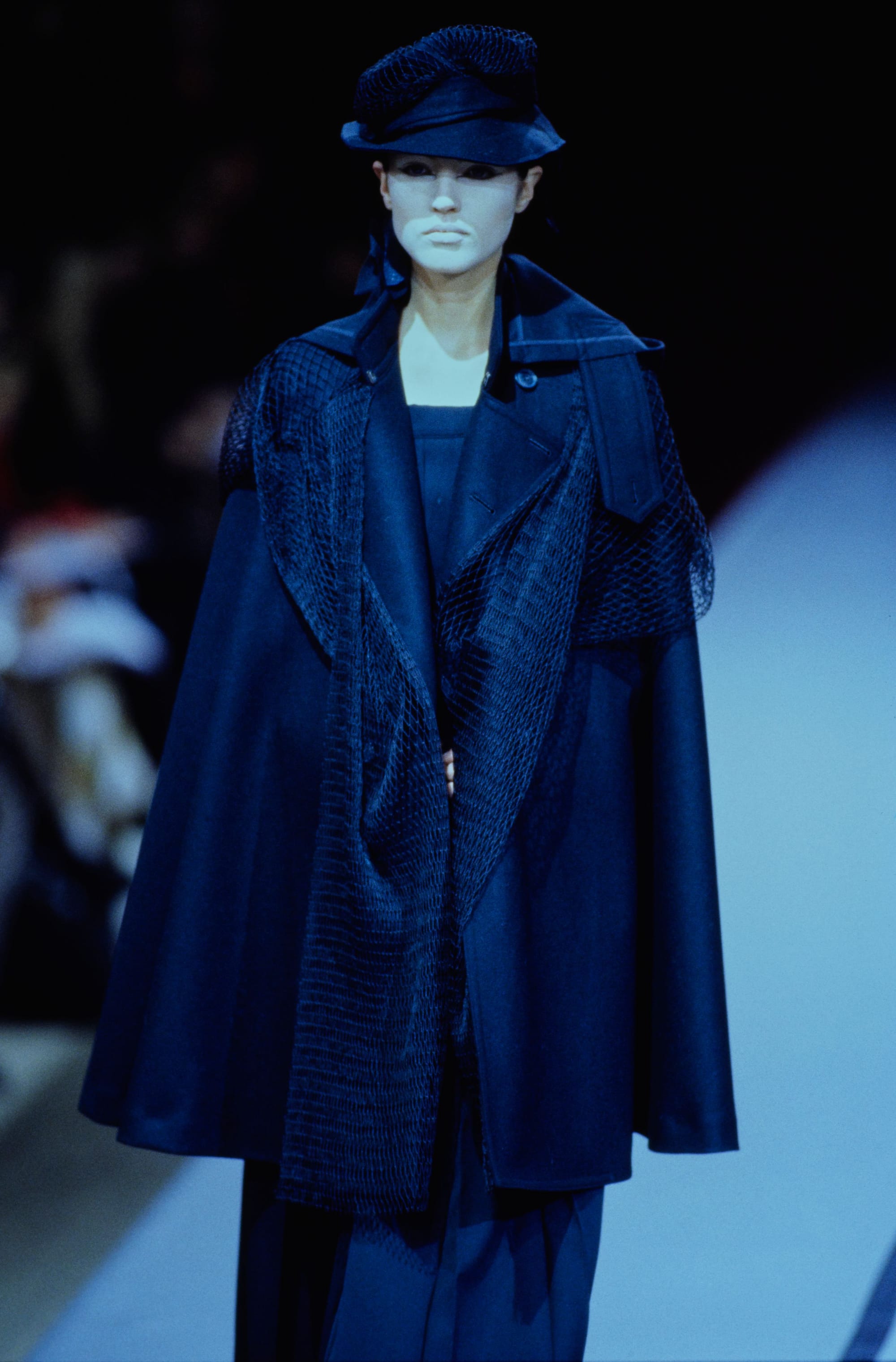 Yohji Yamamoto Fall 1995 detail 4: Navy cape-like coat with mesh front panels, worn with a black sculpted hat.