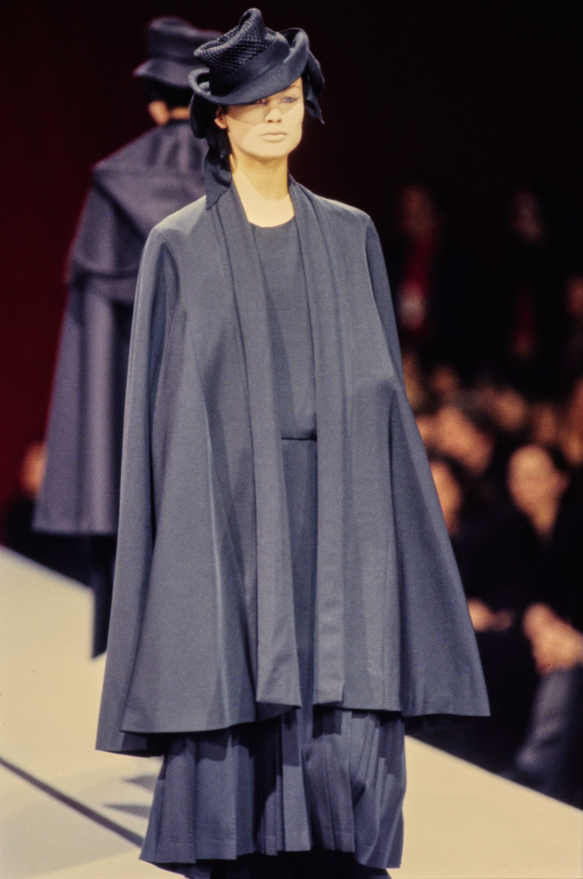 Yohji Yamamoto Fall 1995 detail 5: Slate cape jacket over a pleated dark skirt, topped by a sculptural black hat.