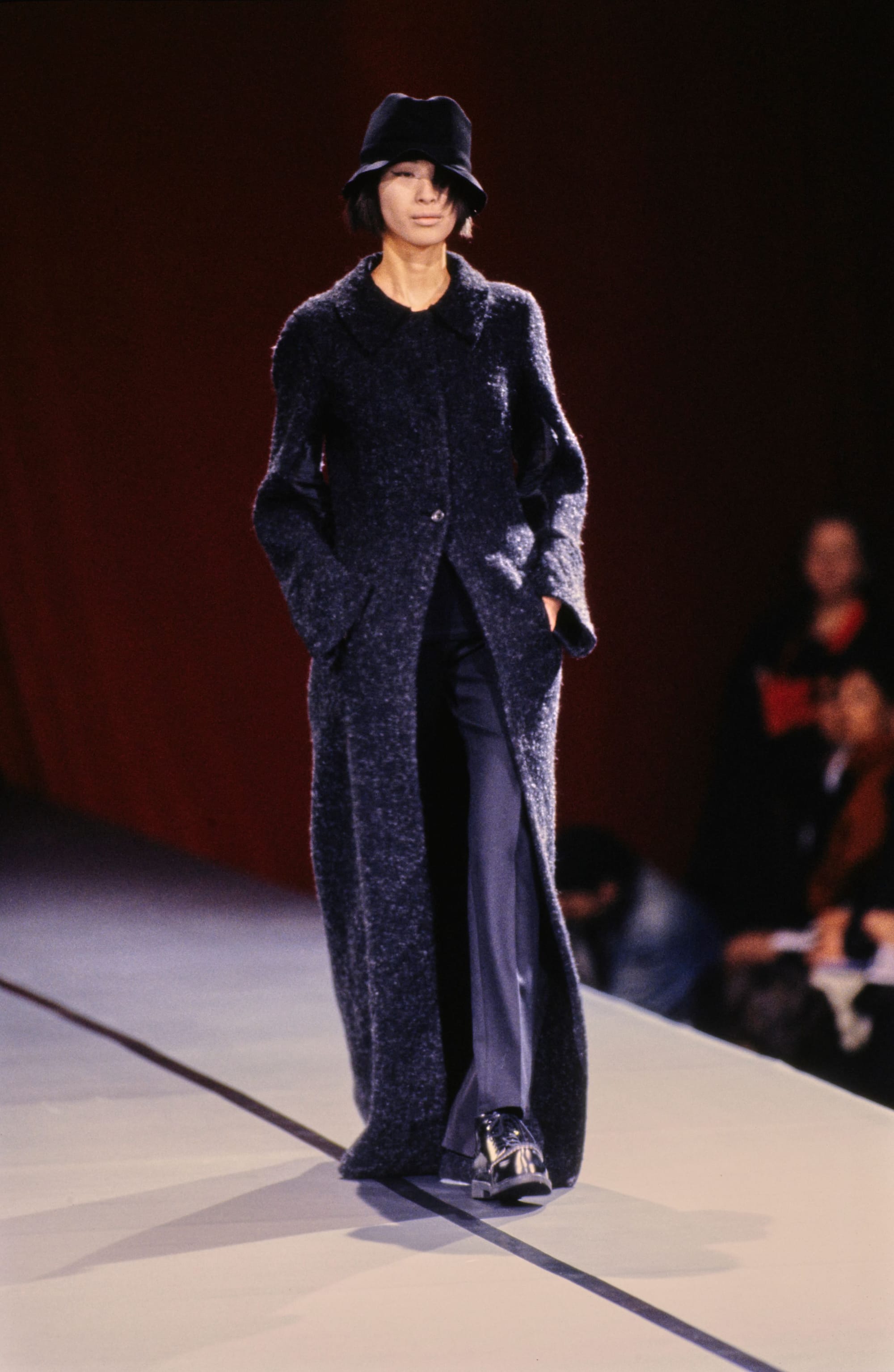 Yohji Yamamoto Fall 1995 detail 6: Full runway view of a dark textured long coat, black hat, wide trousers and glossy shoes.