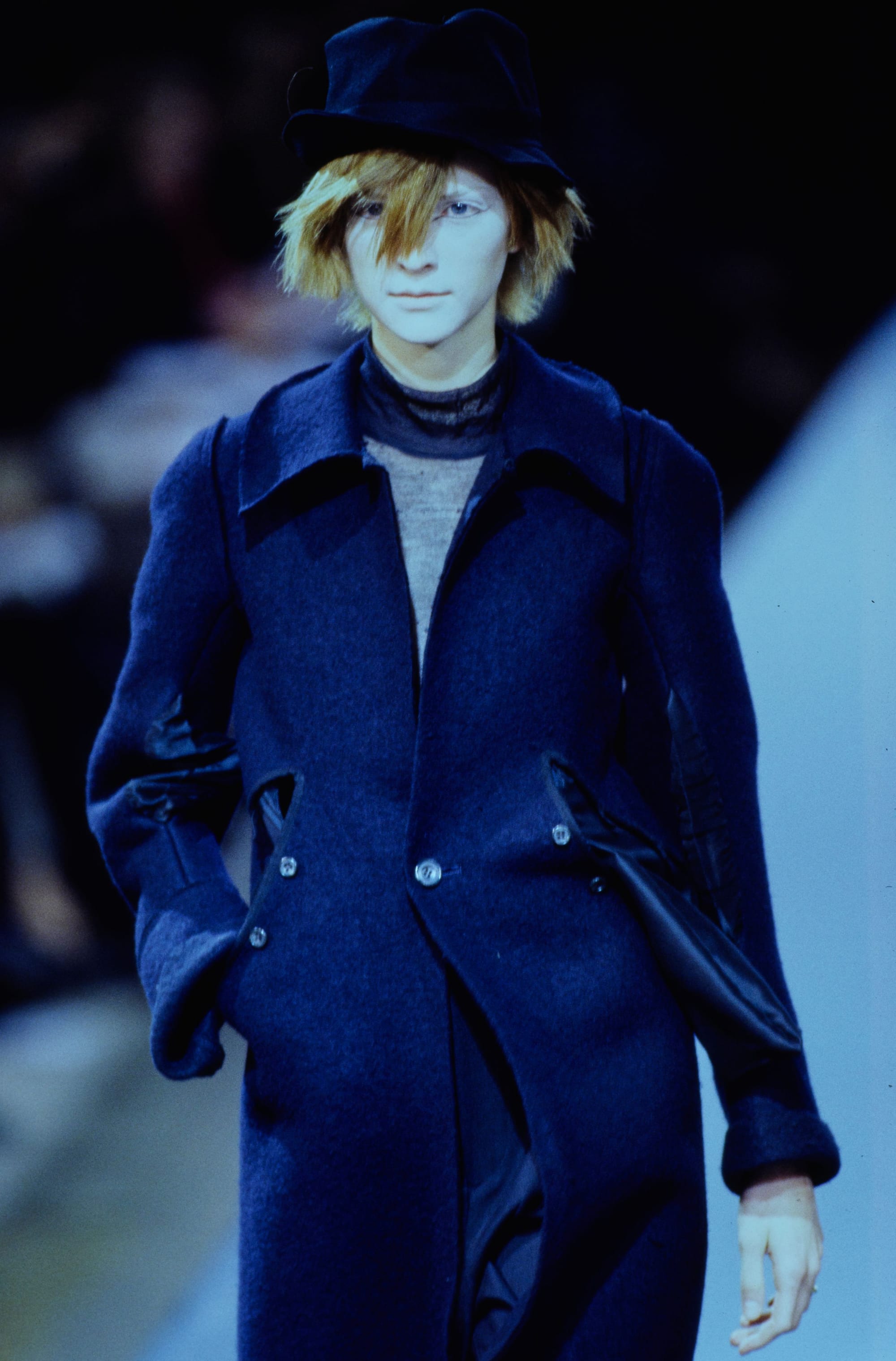 Yohji Yamamoto Fall 1995 detail 7: Navy collared coat with angled waist pockets beneath a black brimmed hat.