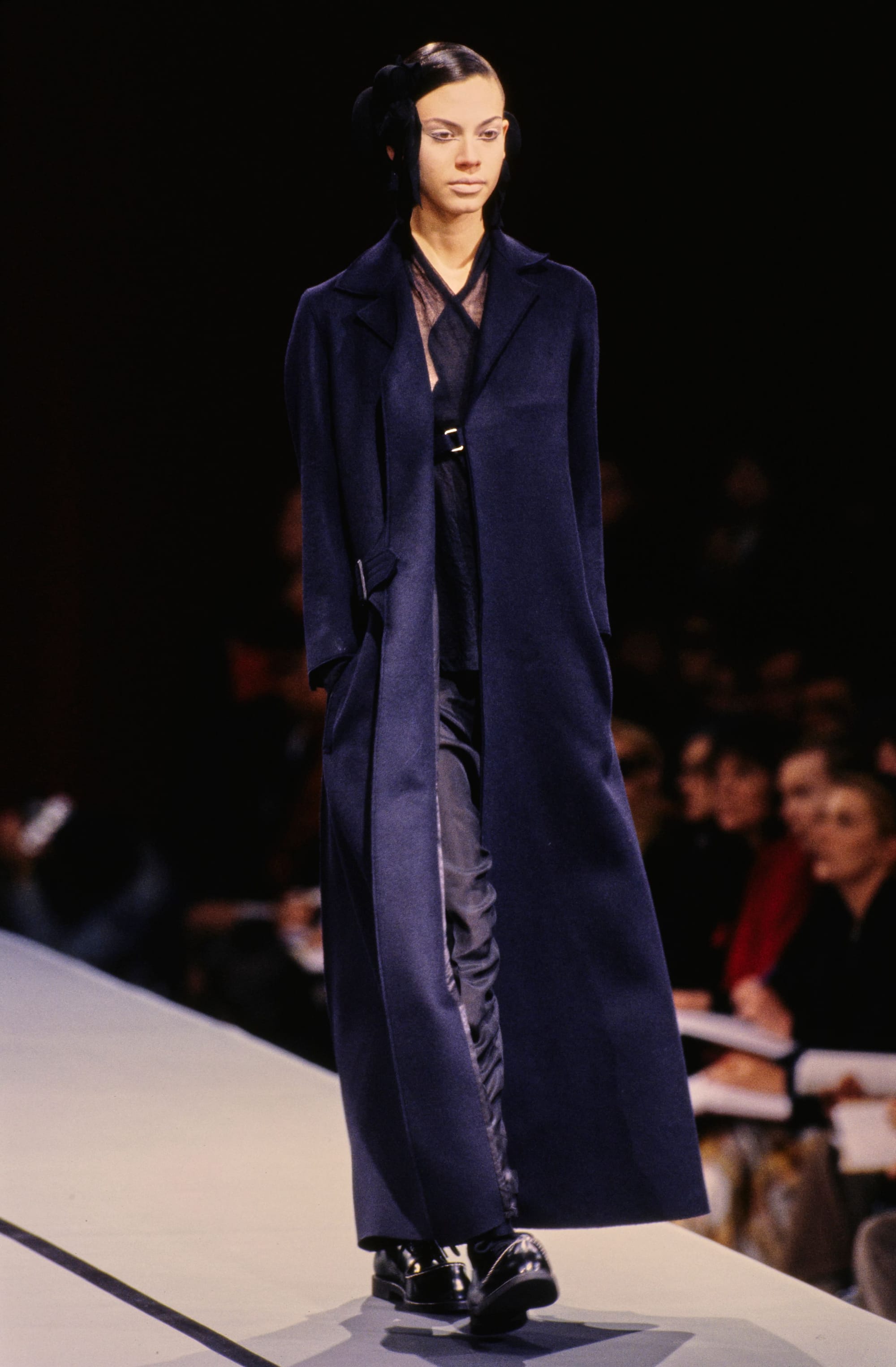 Yohji Yamamoto Fall 1995 detail 11: Long navy coat open over a belted dark layered outfit and black shoes.