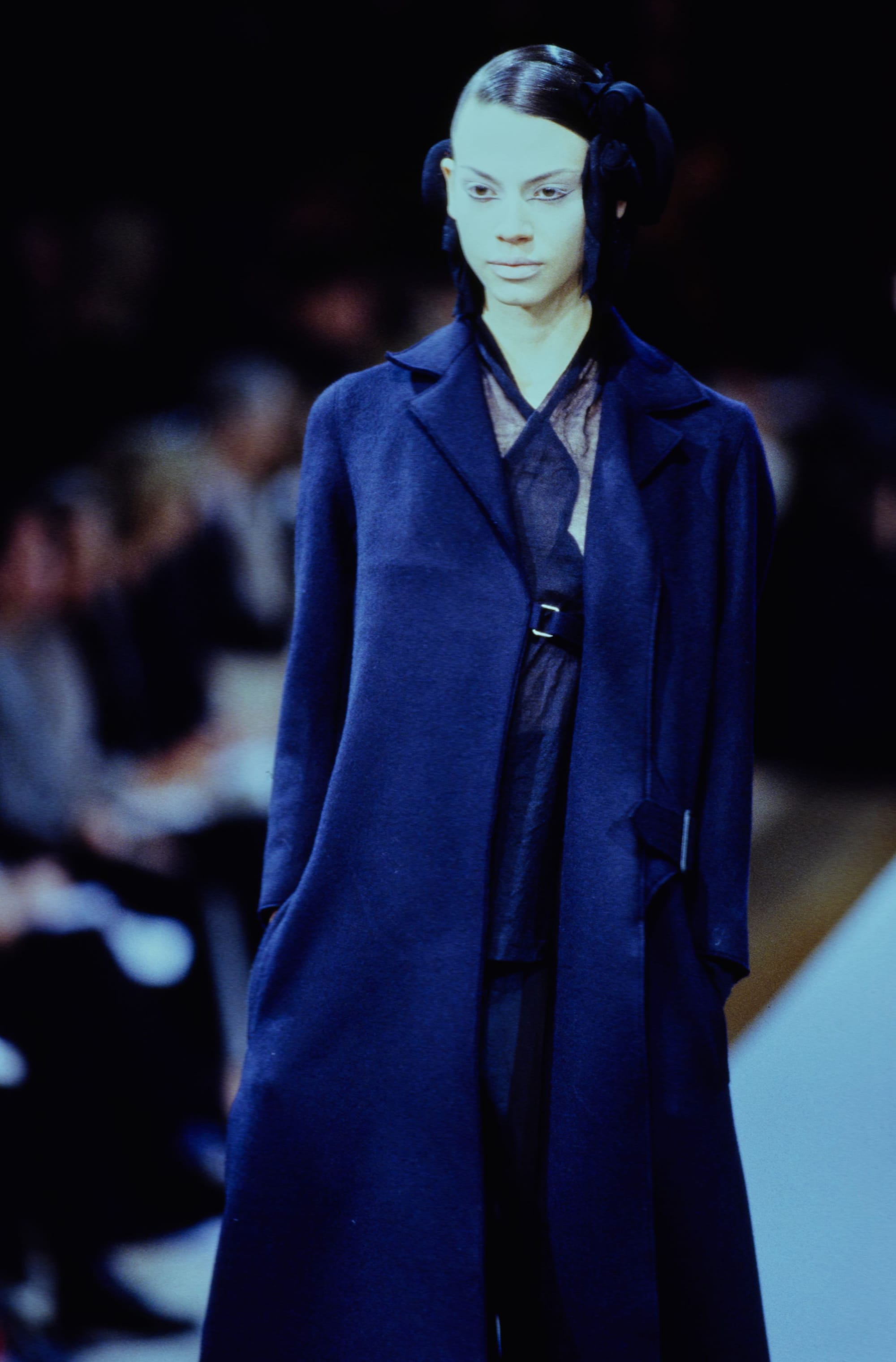 Yohji Yamamoto Fall 1995 detail 12: Close view of a dark coat over a sheer crossed-neck top, with black ribbon hair ornaments.