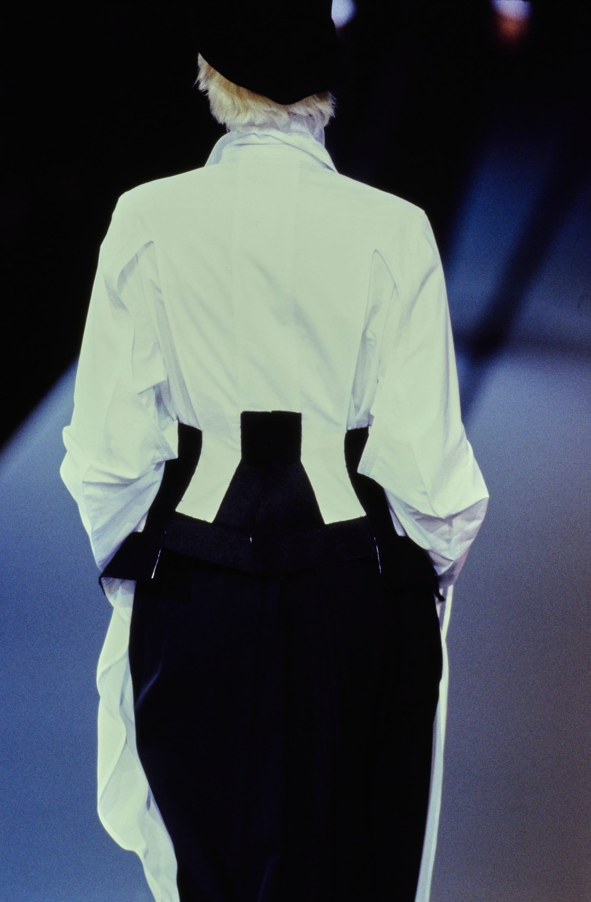 Yohji Yamamoto Fall 1995 detail 14: Rear view of a white blouse shaped by a black waist panel above dark trousers.