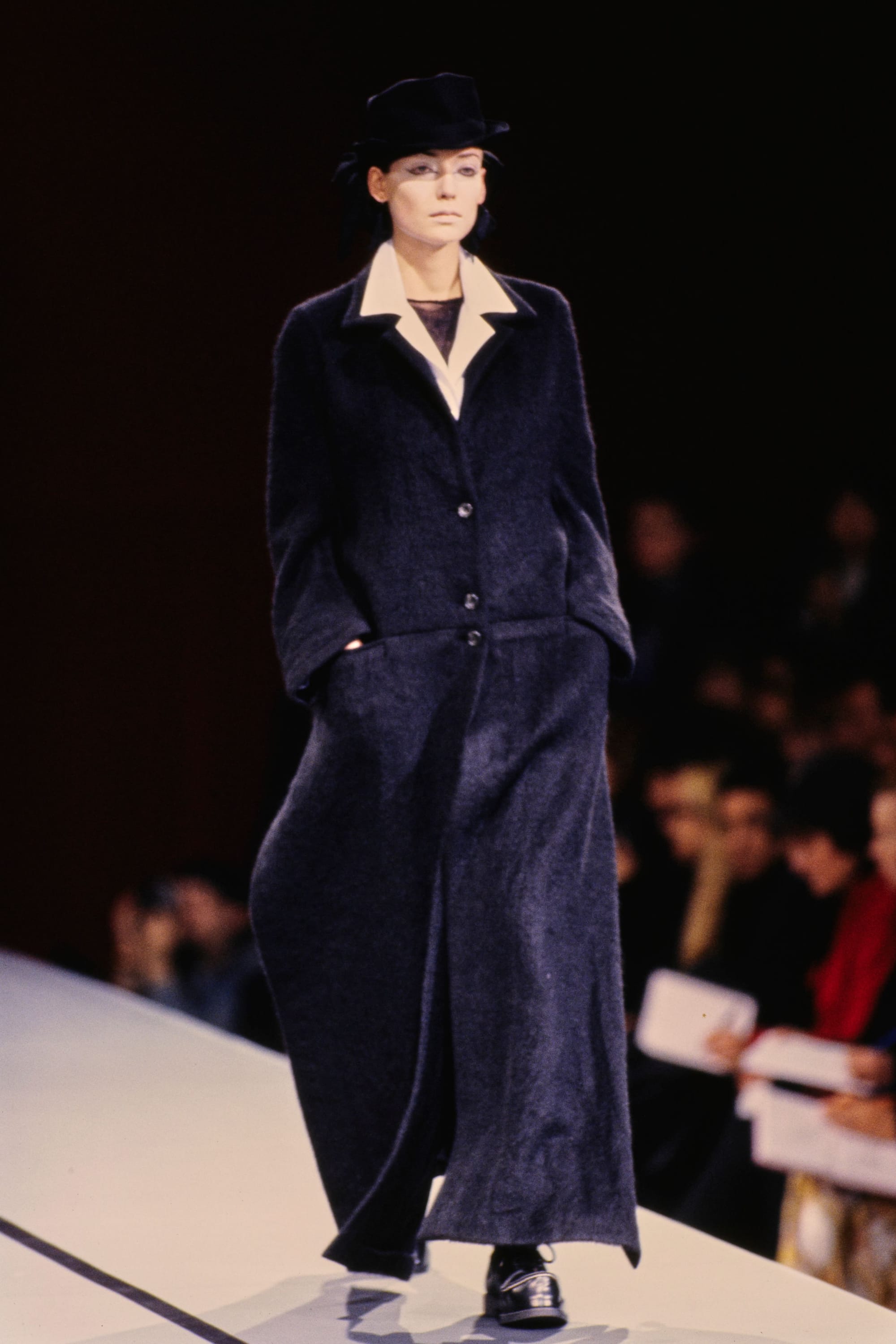 Yohji Yamamoto Fall 1995 detail 15: Long dark coat over a white shirt collar, worn with a black hat.