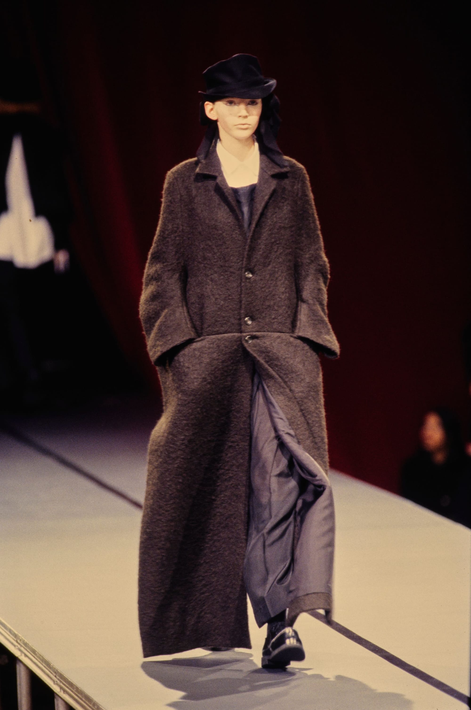 Yohji Yamamoto Fall 1995 detail 16: Long dark brown textured coat over wide grey trousers and a black hat.
