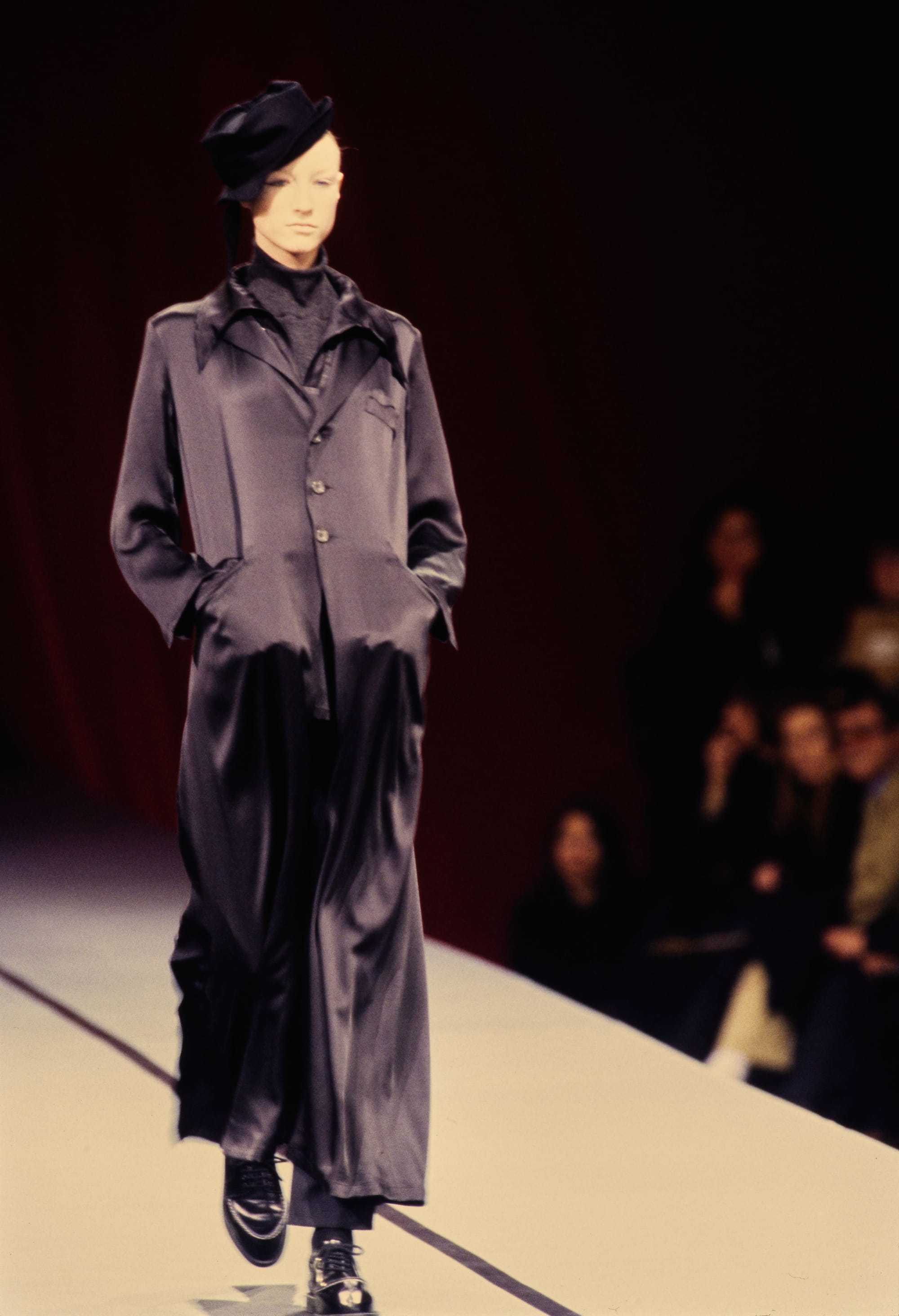 Yohji Yamamoto Fall 1995 detail 20: Long glossy dark coat over a high-neck top, with a tilted black hat.