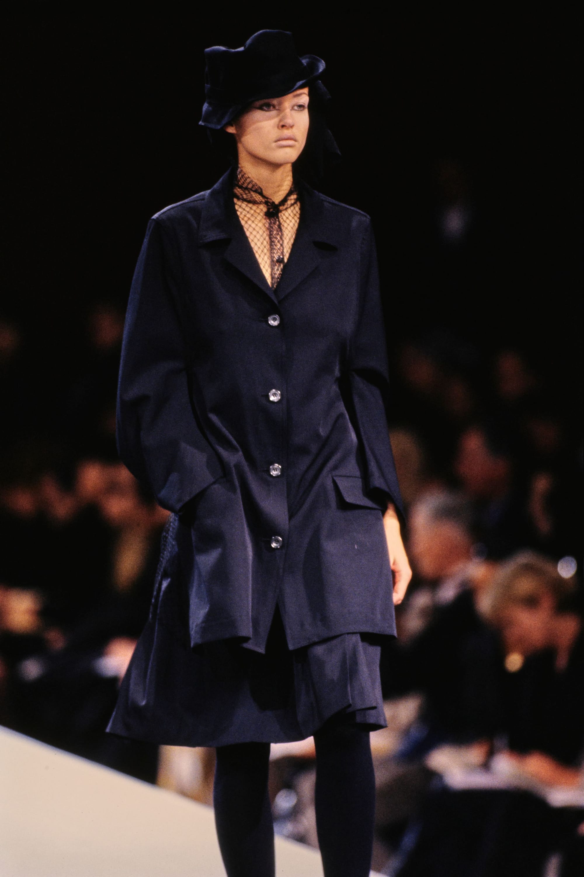 Yohji Yamamoto Fall 1995 detail 22: Navy jacket and short layered skirt over a black fishnet high neck, with a black hat.