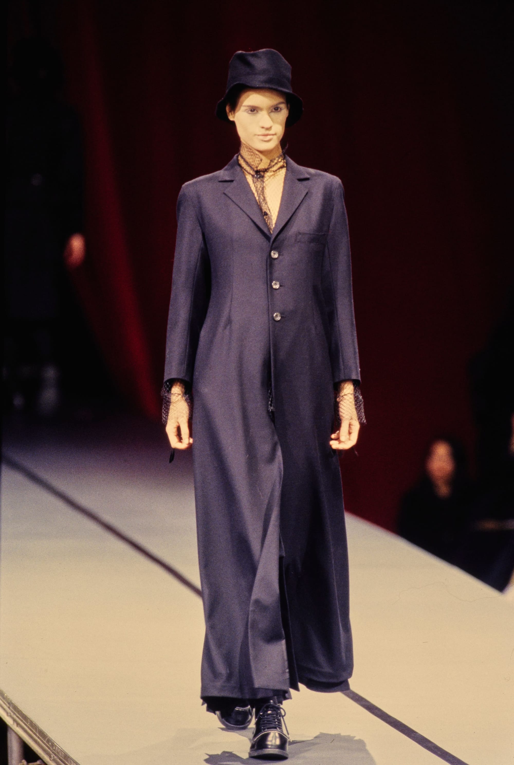Yohji Yamamoto Fall 1995 detail 24: Full navy long coat with fishnet at neck and wrists, worn with a black hat.