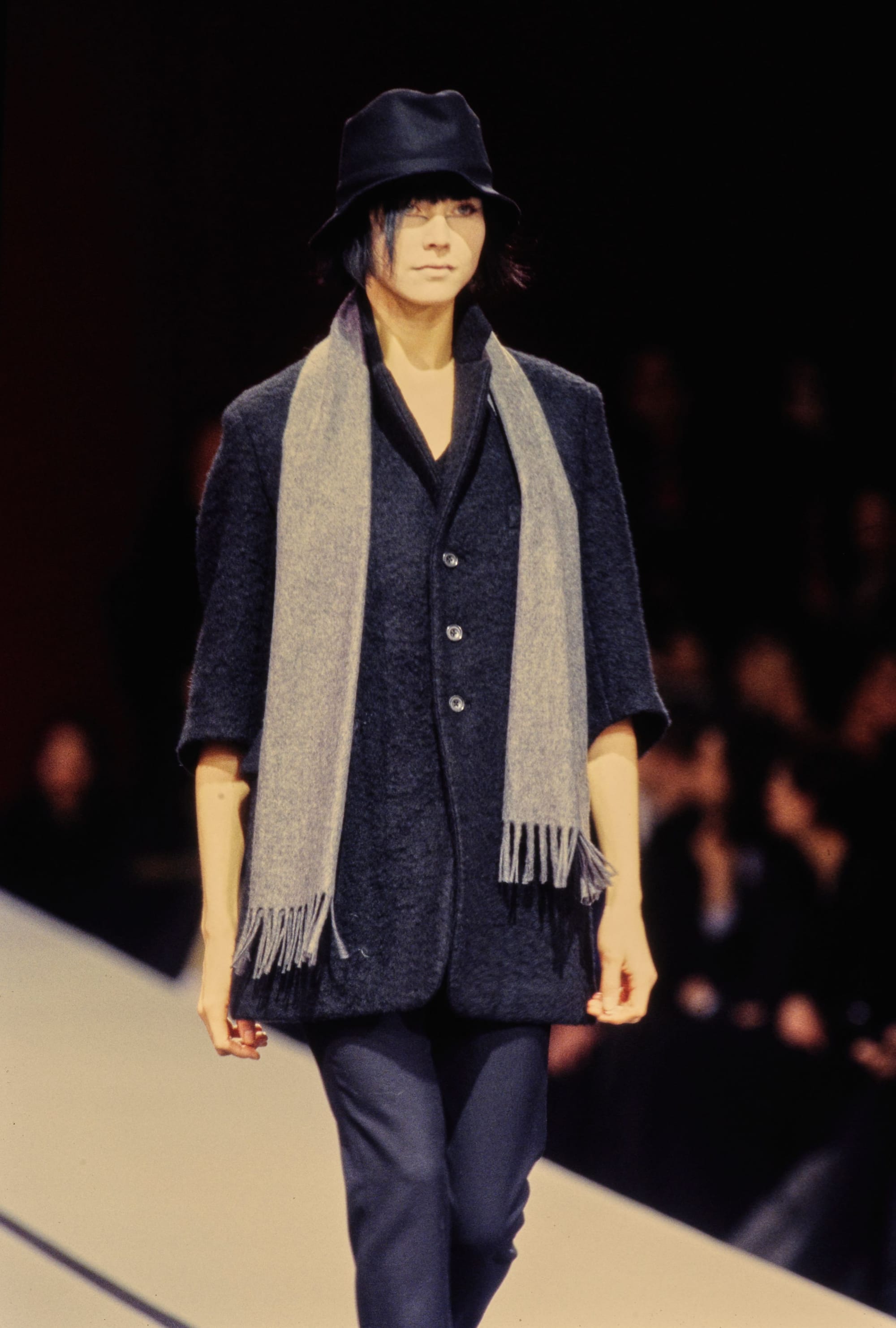 Yohji Yamamoto Fall 1995 detail 28: Black hat, dark short-sleeved jacket and long grey fringed scarf over navy trousers.