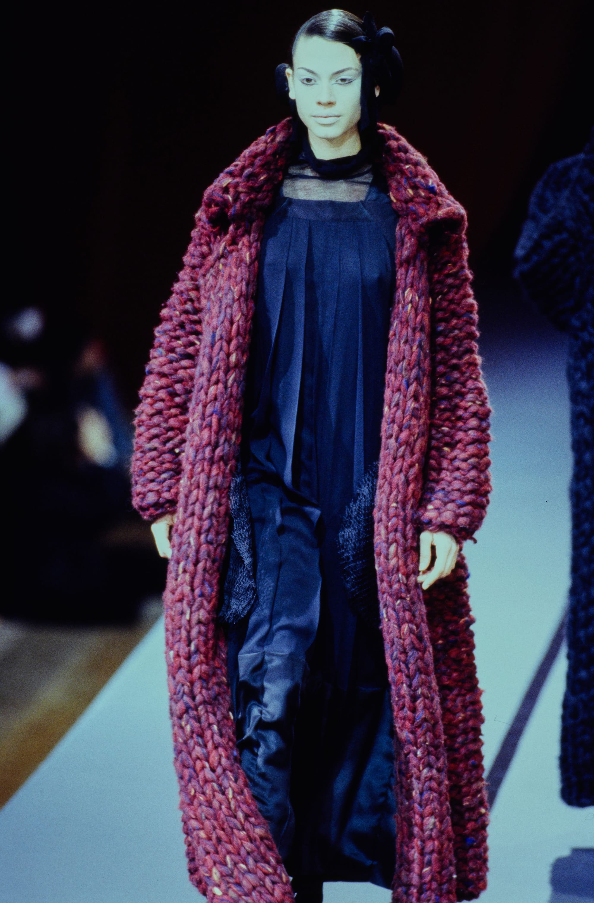 Yohji Yamamoto Fall 1995 detail 34: Full view of a berry-red chunky knit coat around a long navy layered dress.