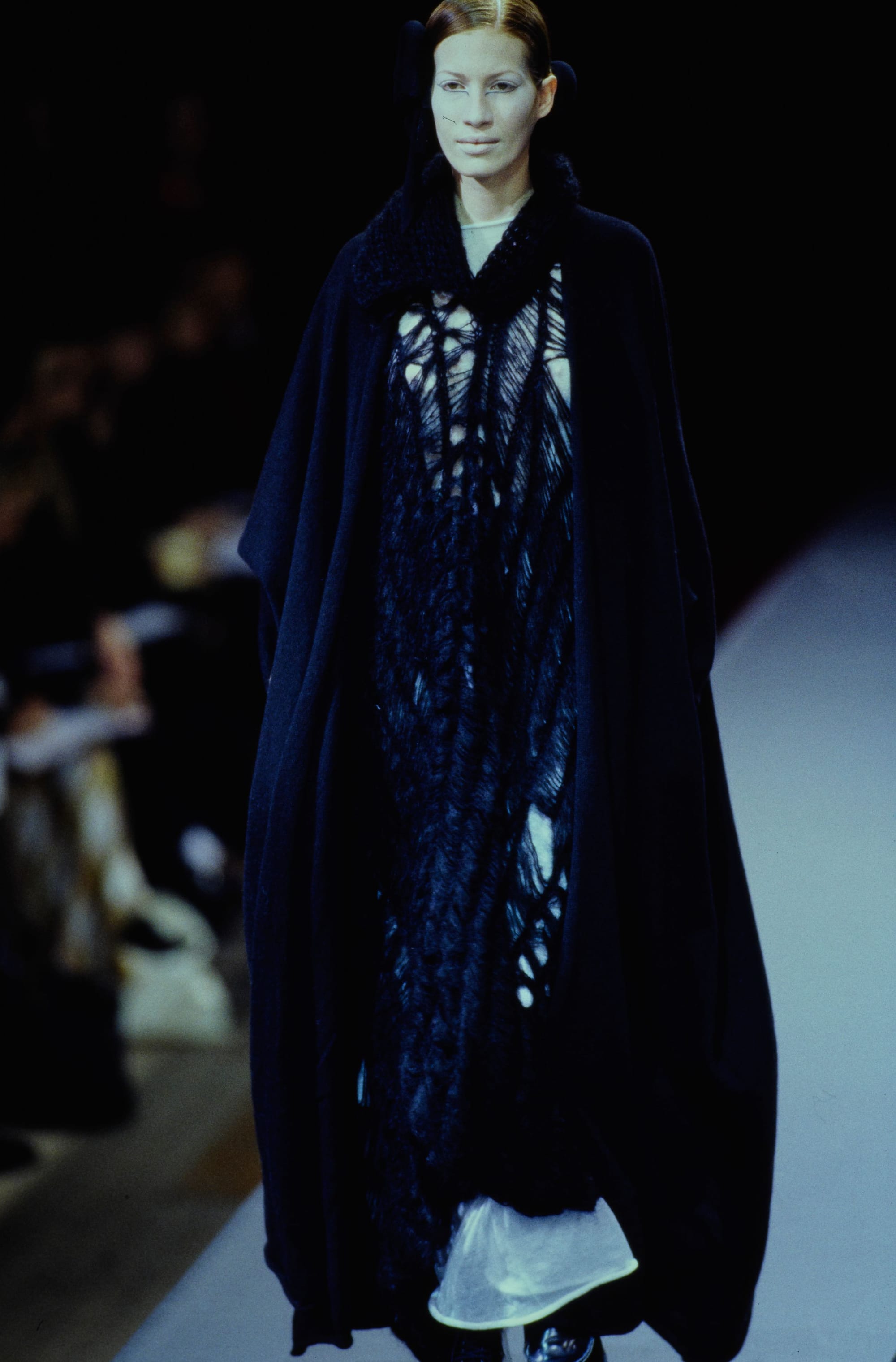 Yohji Yamamoto Fall 1995 detail 38: Black openwork dress under a flowing dark cape, with a white layer visible at the hem.