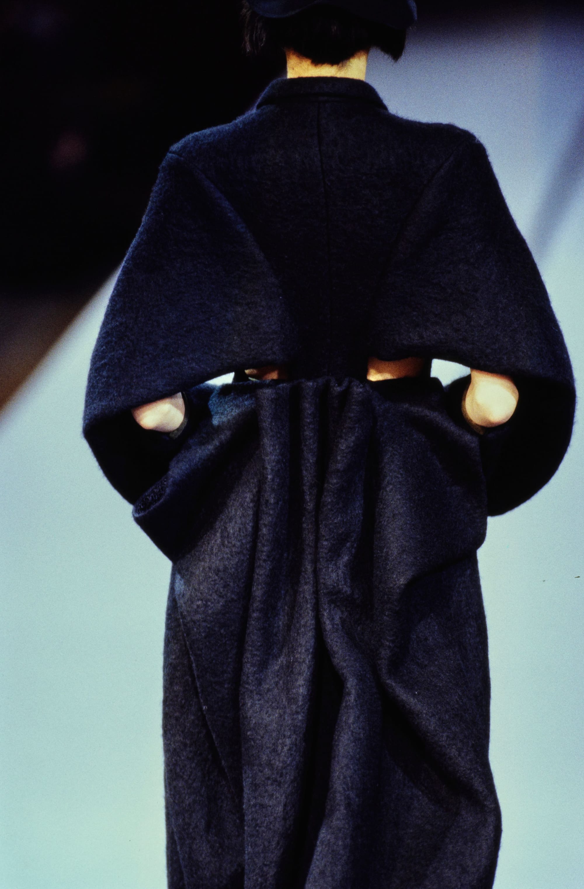 Yohji Yamamoto Fall 1995 detail 58: Rear view of a dark coat with broad cape sleeves and fabric gathered at the waist.