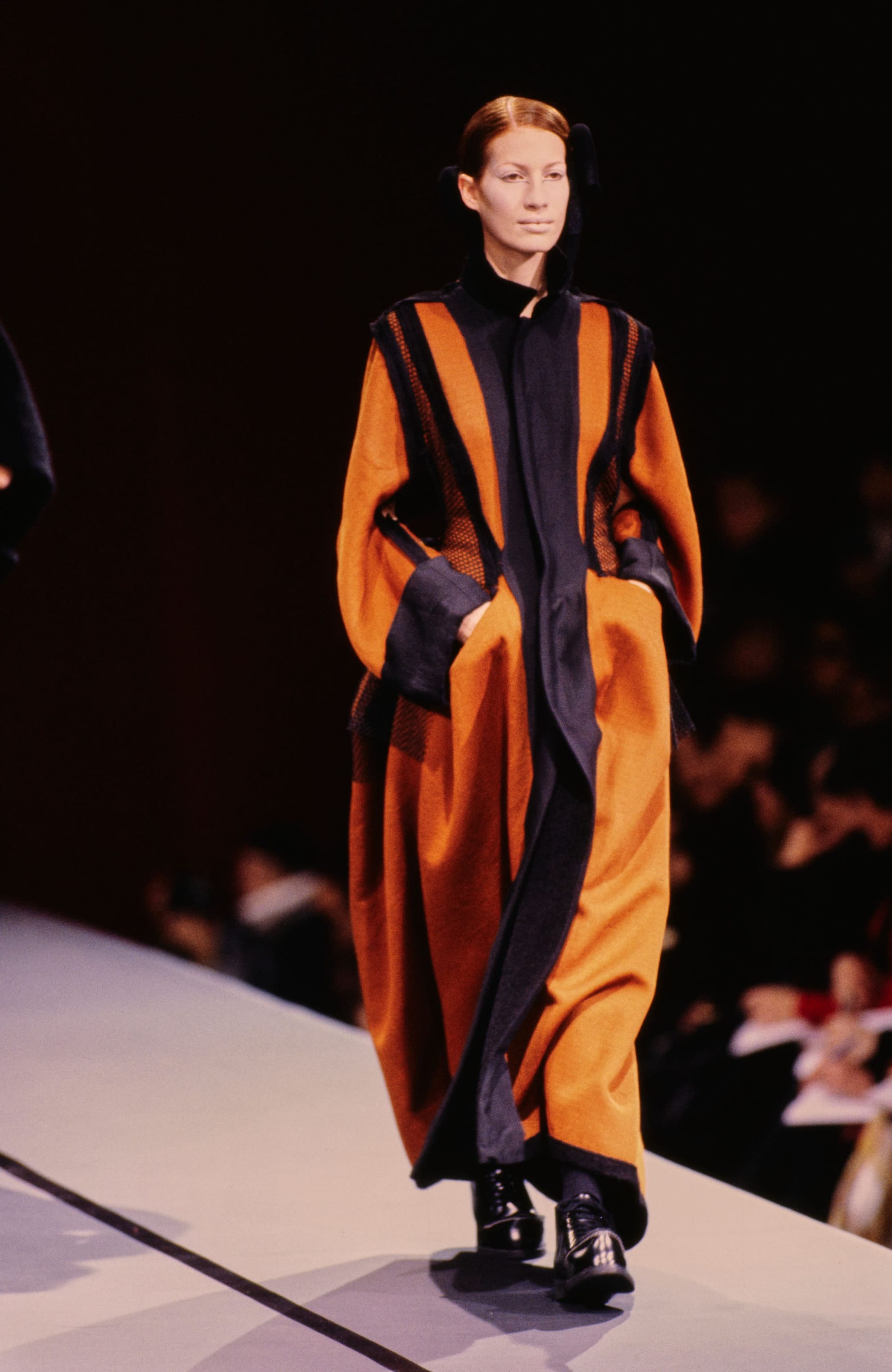 Yohji Yamamoto Fall 1995 detail 59: Long mustard and navy coat with contrasting front panels and black mesh at the sides.