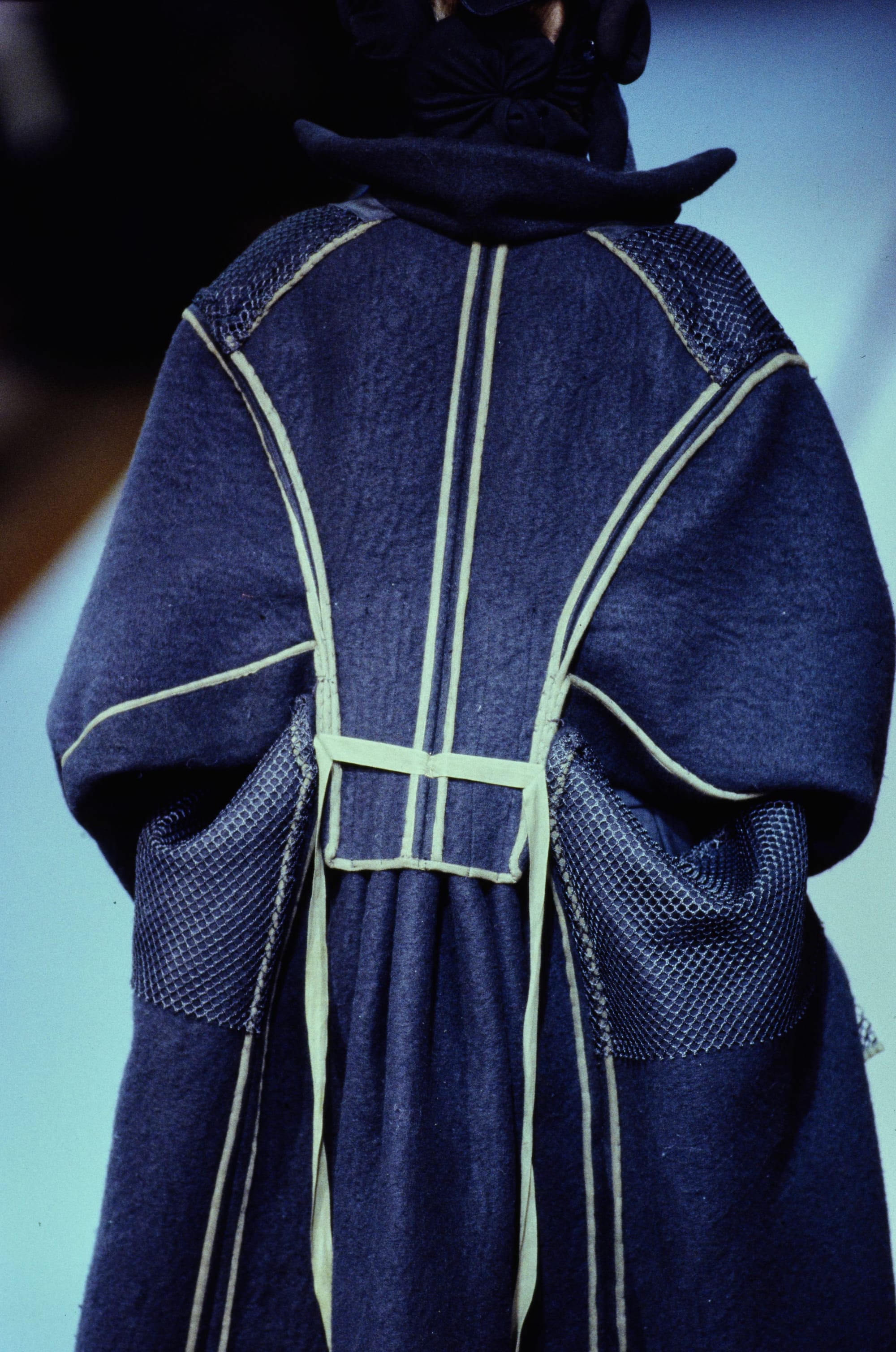Yohji Yamamoto Fall 1995 detail 64: Rear navy coat with pale piping and black mesh panels projecting at the hips.