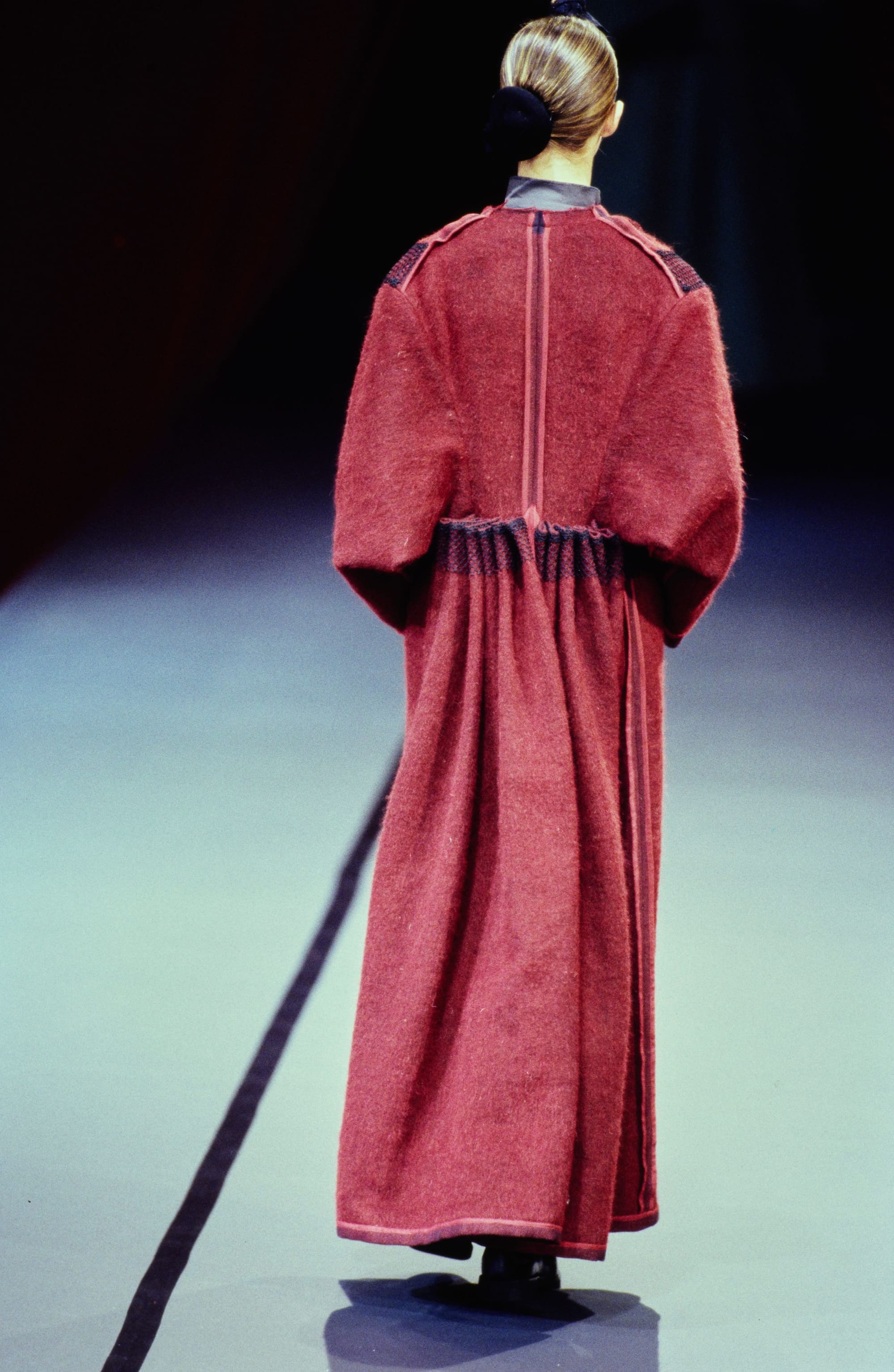 Yohji Yamamoto Fall 1995 detail 65: Rear view of a muted red long coat with a gathered waist and vertical back seams.