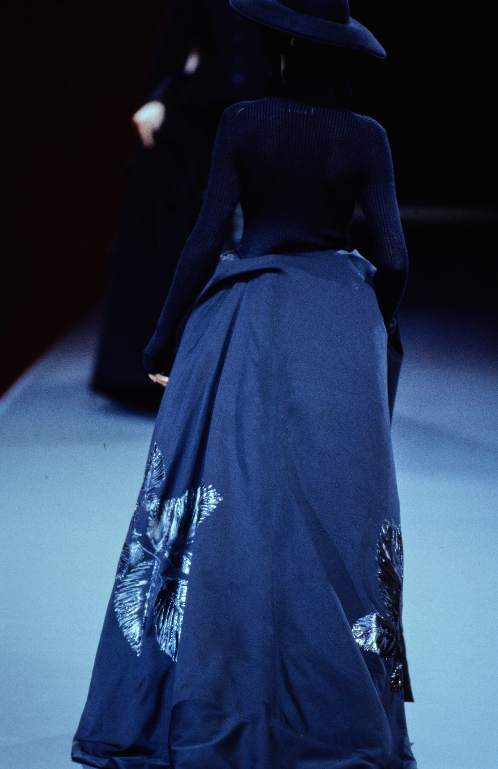 Yohji Yamamoto Fall 1995 detail 69: Rear view of a ribbed navy top and blue skirt with shiny motifs near the hem.