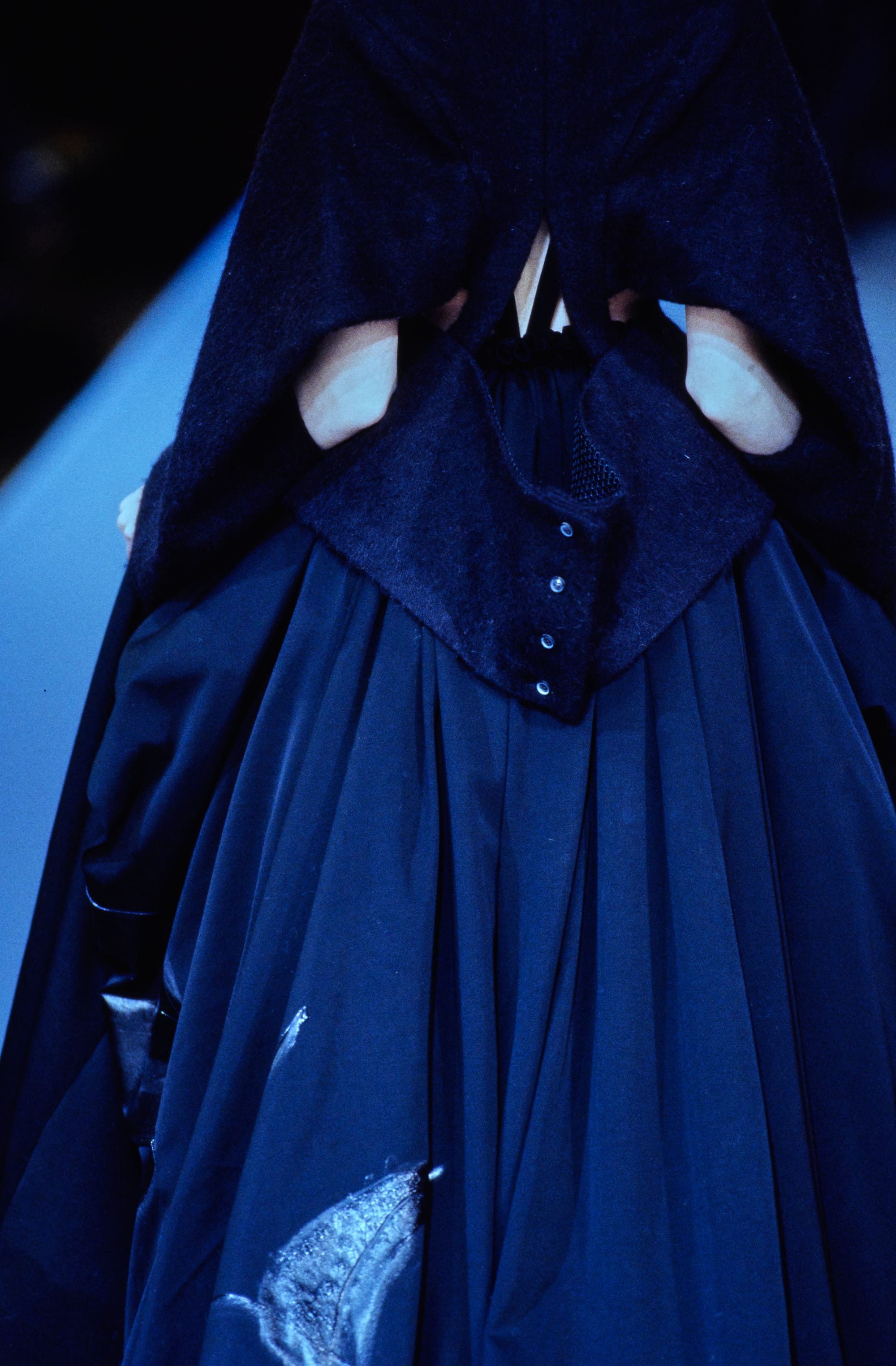 Yohji Yamamoto Fall 1995 detail 71: Rear dark cape with waist openings and a buttoned flap above a pleated skirt.