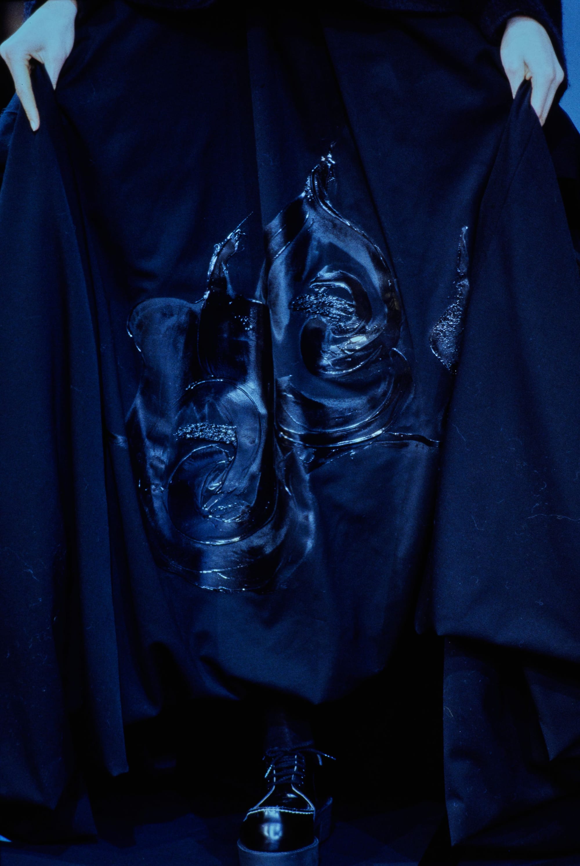 Yohji Yamamoto Fall 1995 detail 72: Close view of a dark skirt held open to show glossy raised motifs and a black shoe.