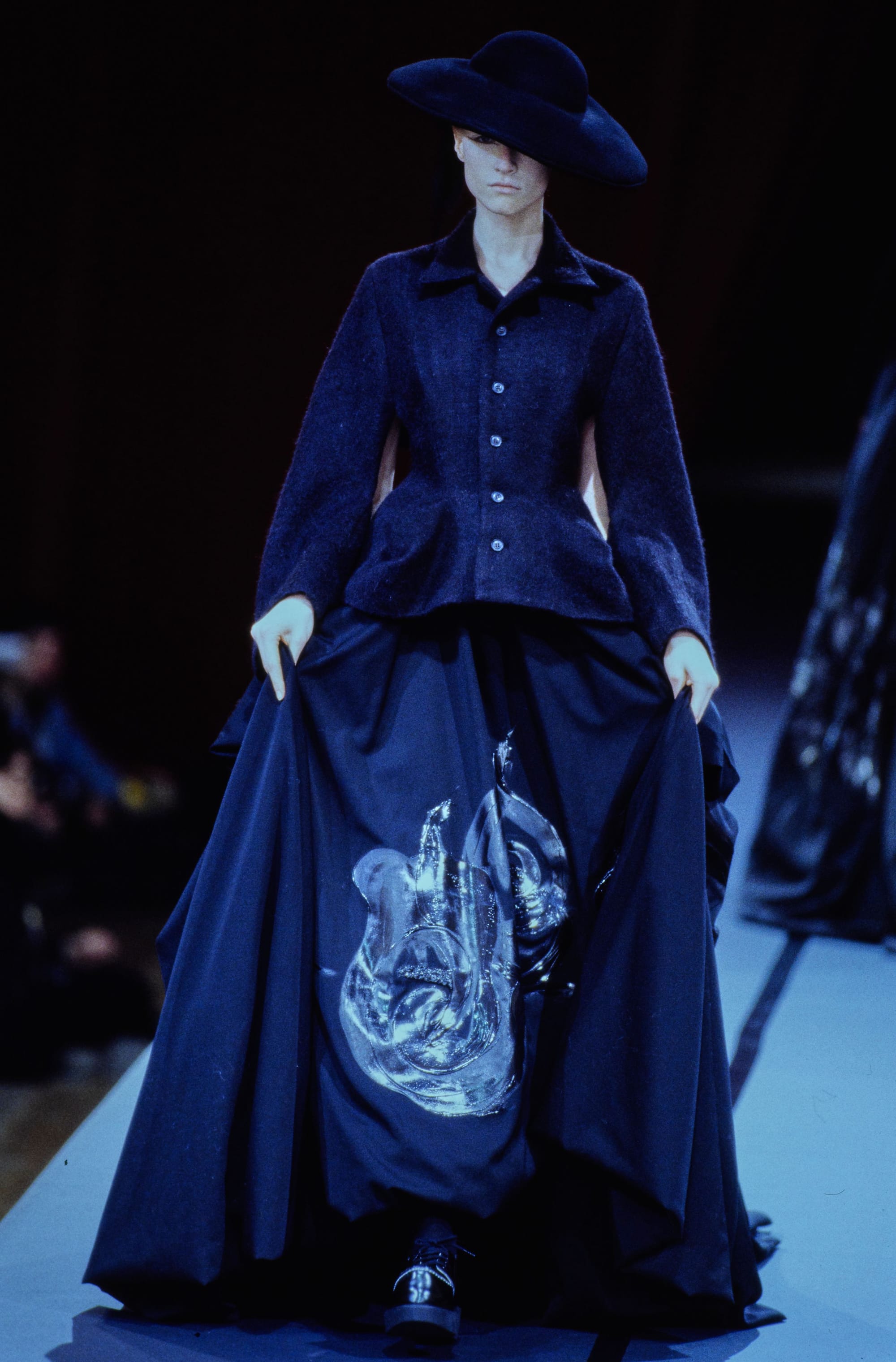 Yohji Yamamoto Fall 1995 detail 73: Broad black hat and fitted navy jacket above a full skirt with shiny motifs.