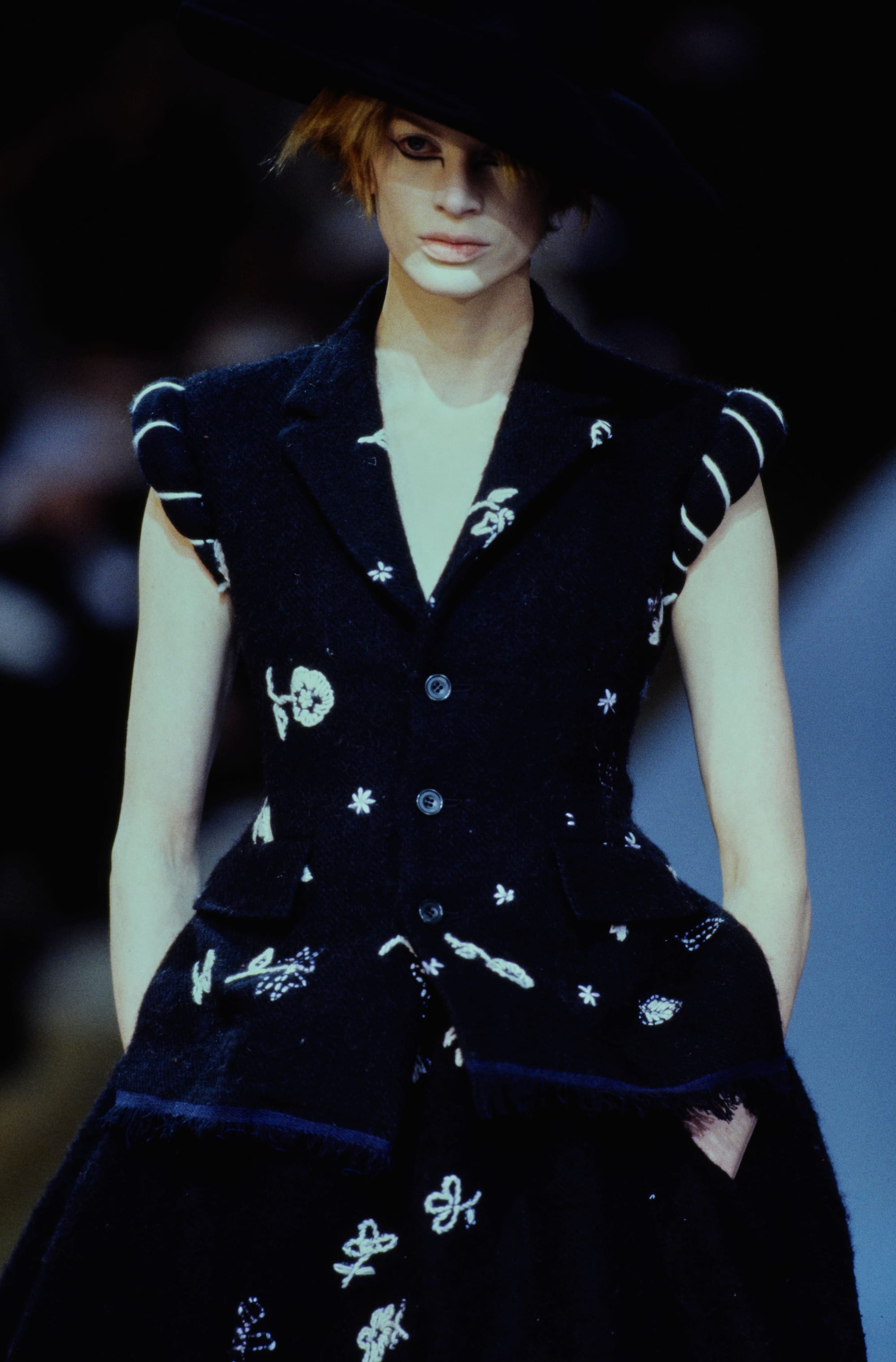 Yohji Yamamoto Fall 1995 detail 74: Black sleeveless vest with small white floral embroidery and rounded shoulders beneath a broad hat.