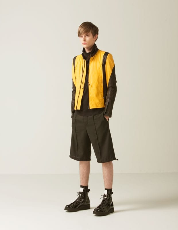Male model in a yellow sleeveless Man of Moods zip vest, black high-neck top, wide black shorts, and boots.
