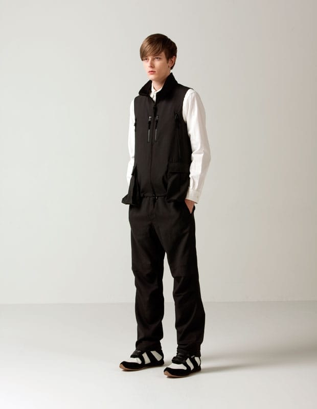 Male model in a black Man of Moods zip utility vest over a white shirt with black trousers.