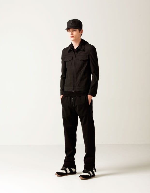 Male model in a black Man of Moods zip-front work jacket layered over a hooded top with trousers and cap.