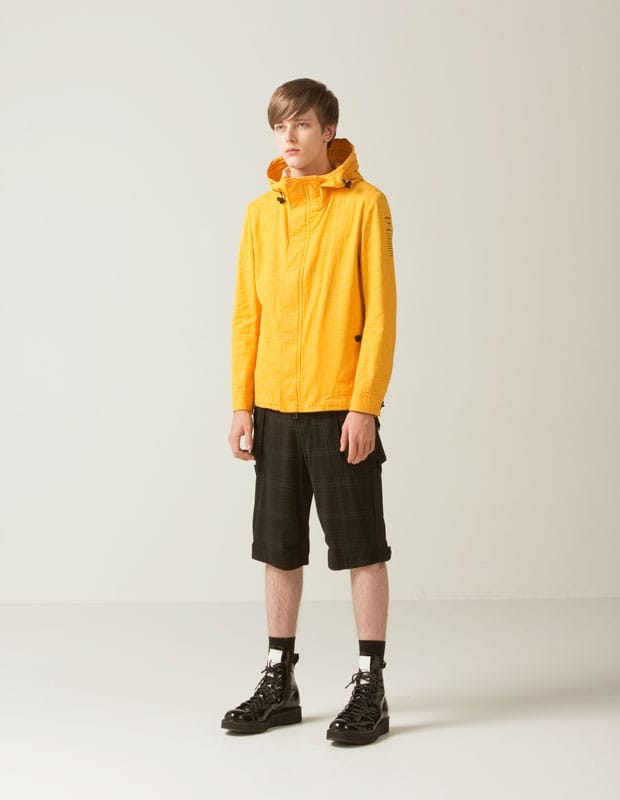 Male model in a bright yellow Man of Moods hooded jacket with dark plaid knee-length shorts.