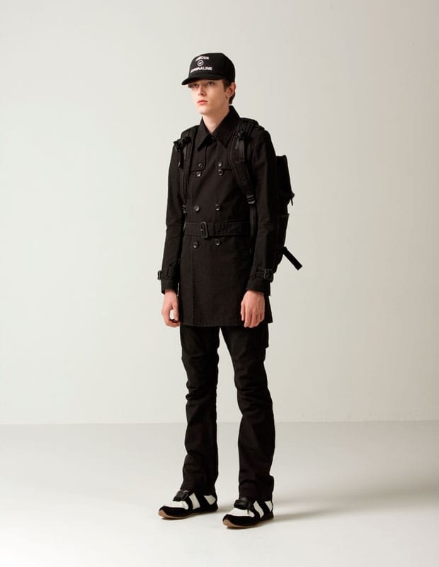 Male model in a black belted double-breasted Man of Moods coat with black trousers, backpack, and cap.