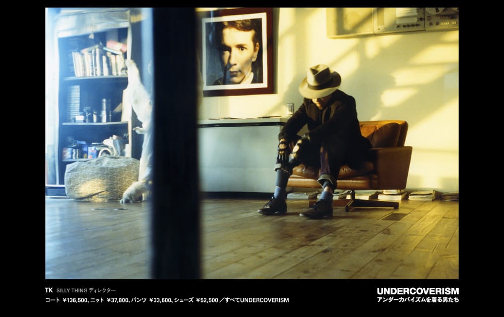 A man in a dark coat and brimmed hat seated on a brown sofa in a warm interior.