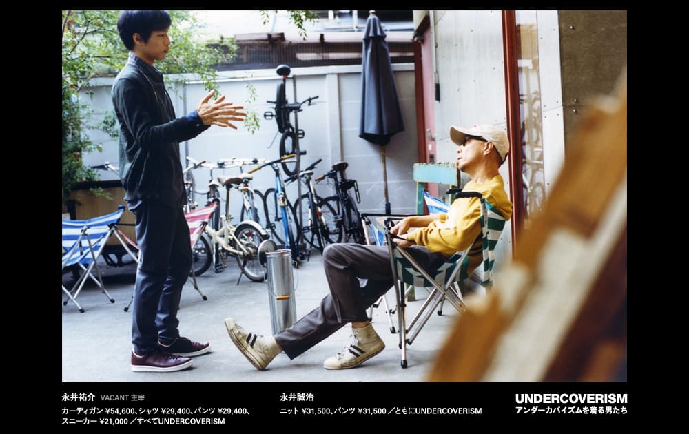 Two men in a courtyard, one standing in a dark cardigan and the other seated in a mustard sweatshirt and cap.