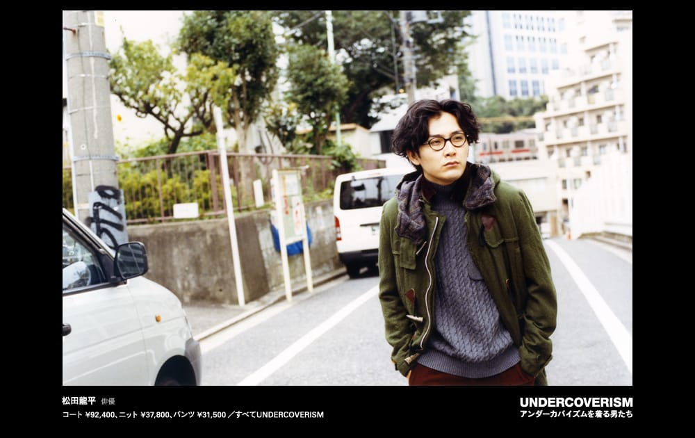A man with round glasses wearing an olive coat and textured grey sweater on a city street.