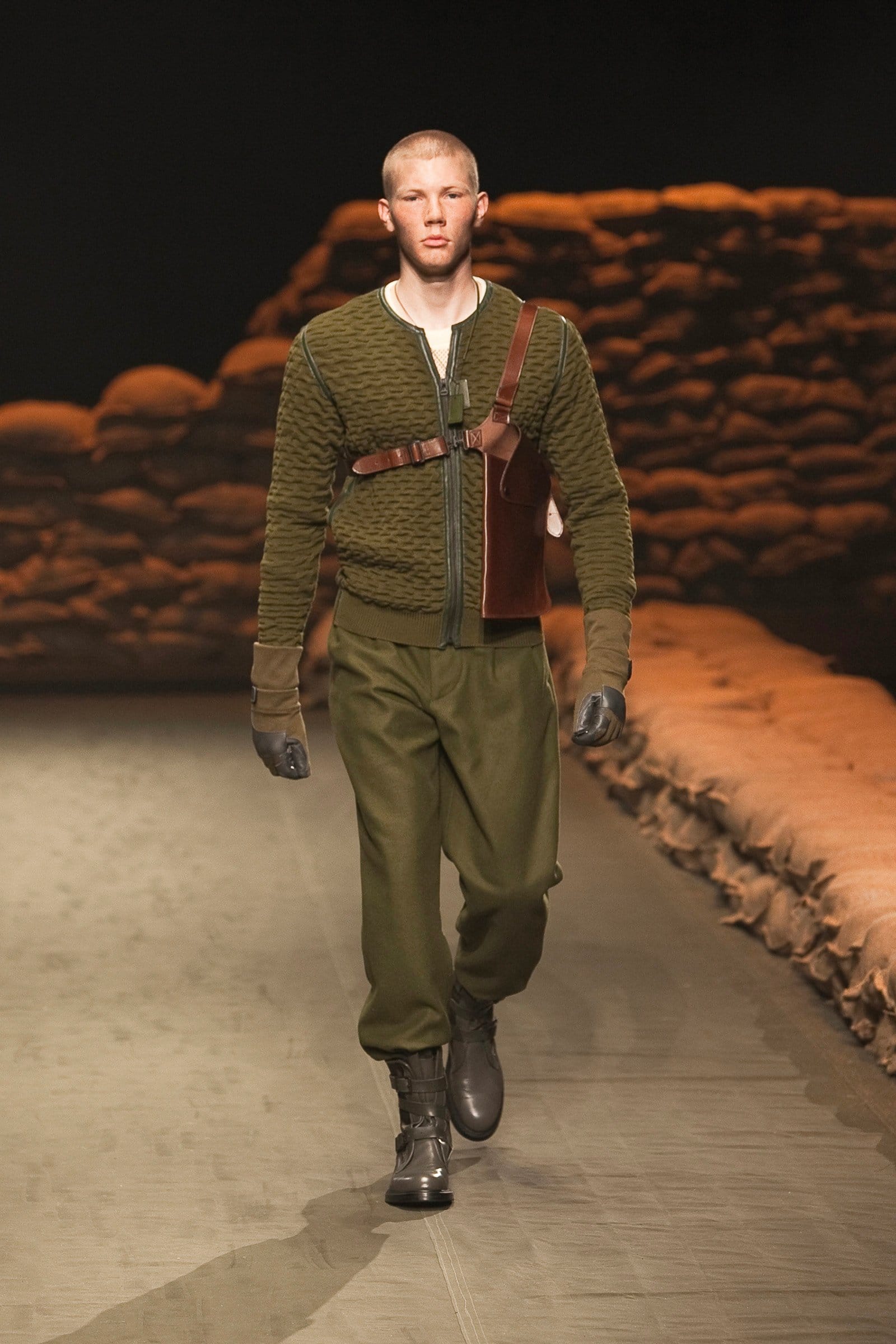 N.Hoolywood Autumn/Winter 2010–11 runway look 4: olive textured cardigan with loose trousers, leather chest harness, gloves, and boots.