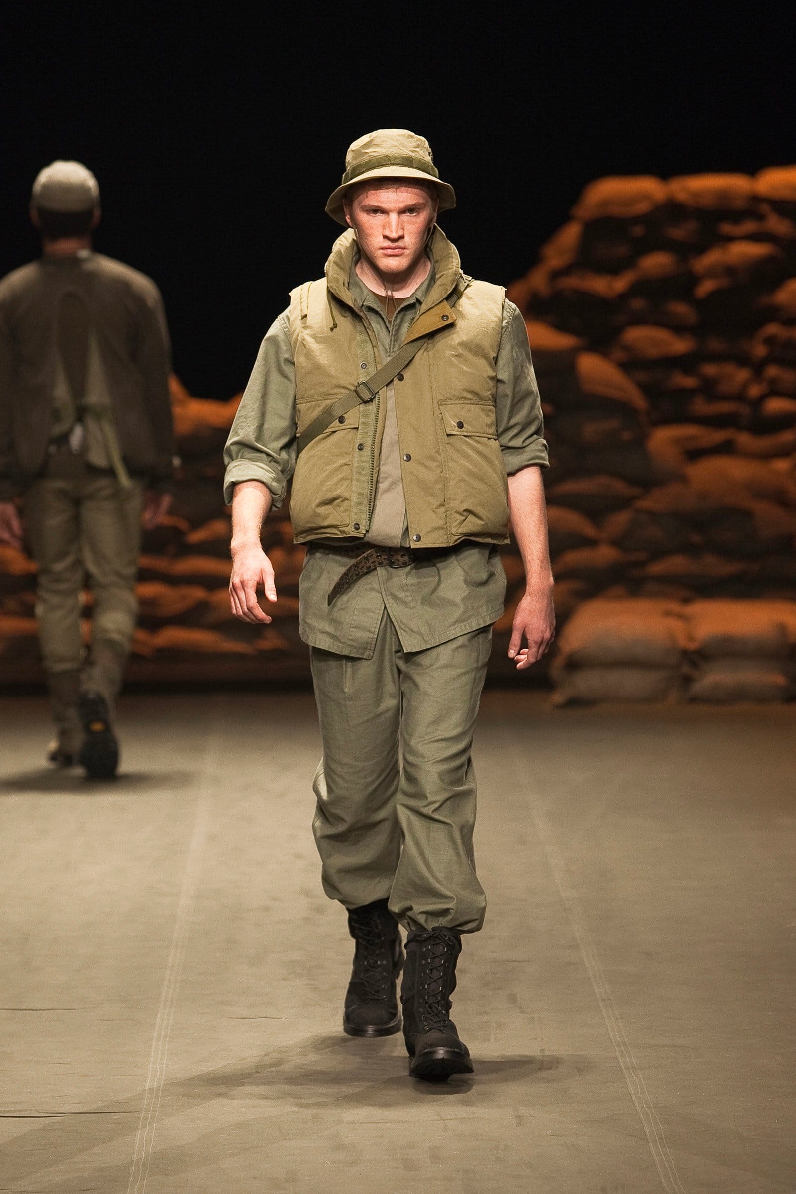 N.Hoolywood Autumn/Winter 2010–11 runway look 10: khaki hooded utility vest over field layers with cargo trousers and bucket hat.