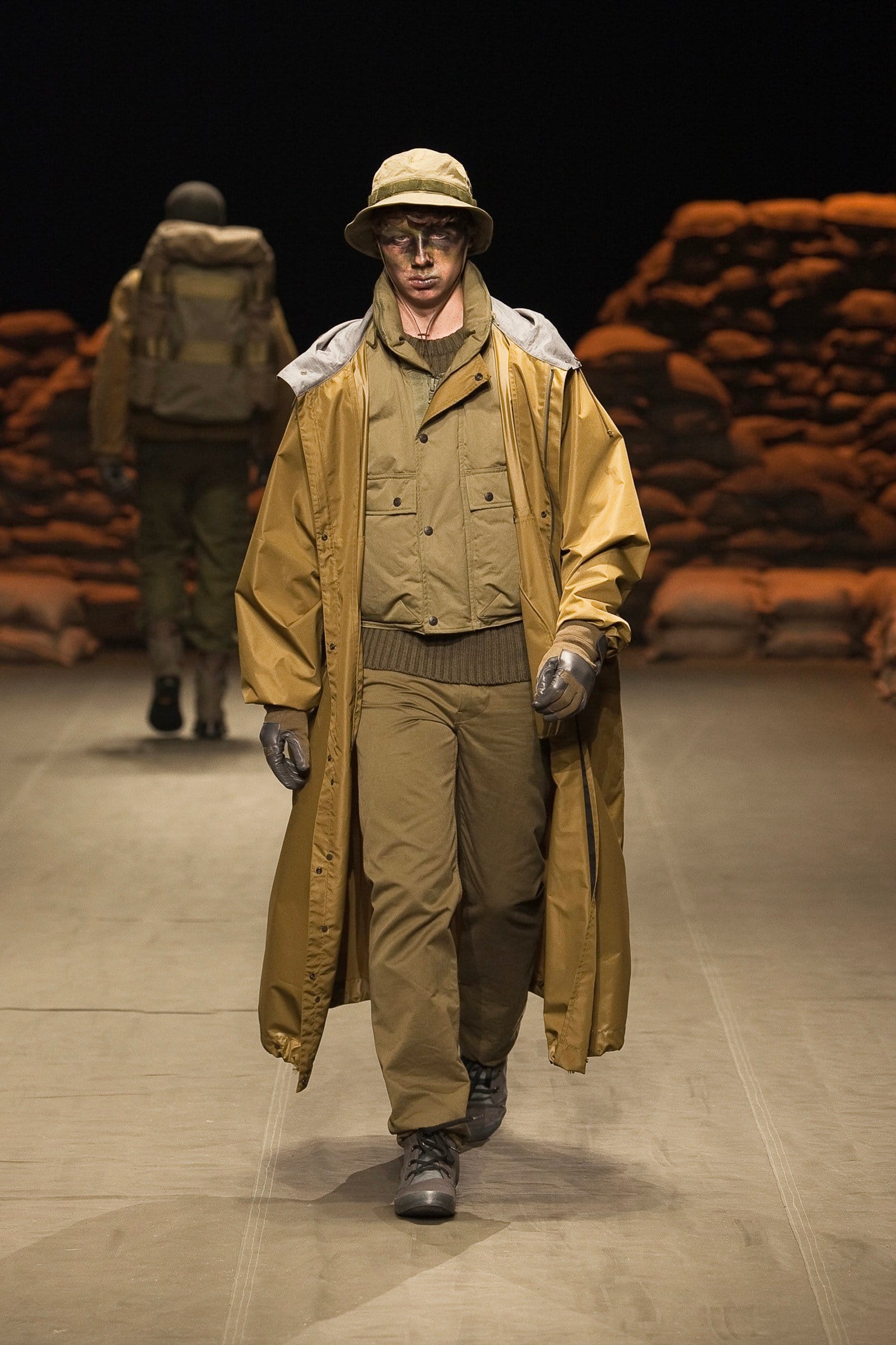 N.Hoolywood Autumn/Winter 2010–11 runway look 12: long mustard raincoat over layered khaki military pieces with bucket hat.