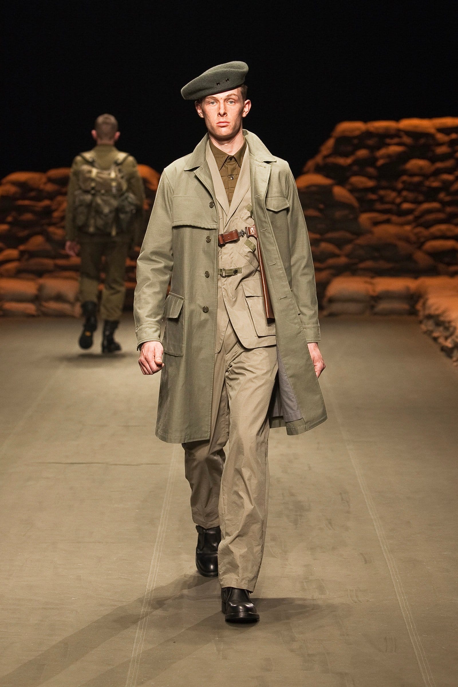 N.Hoolywood Autumn/Winter 2010–11 runway look 22: pale khaki belted trench coat with chest harness, matching trousers, and beret.