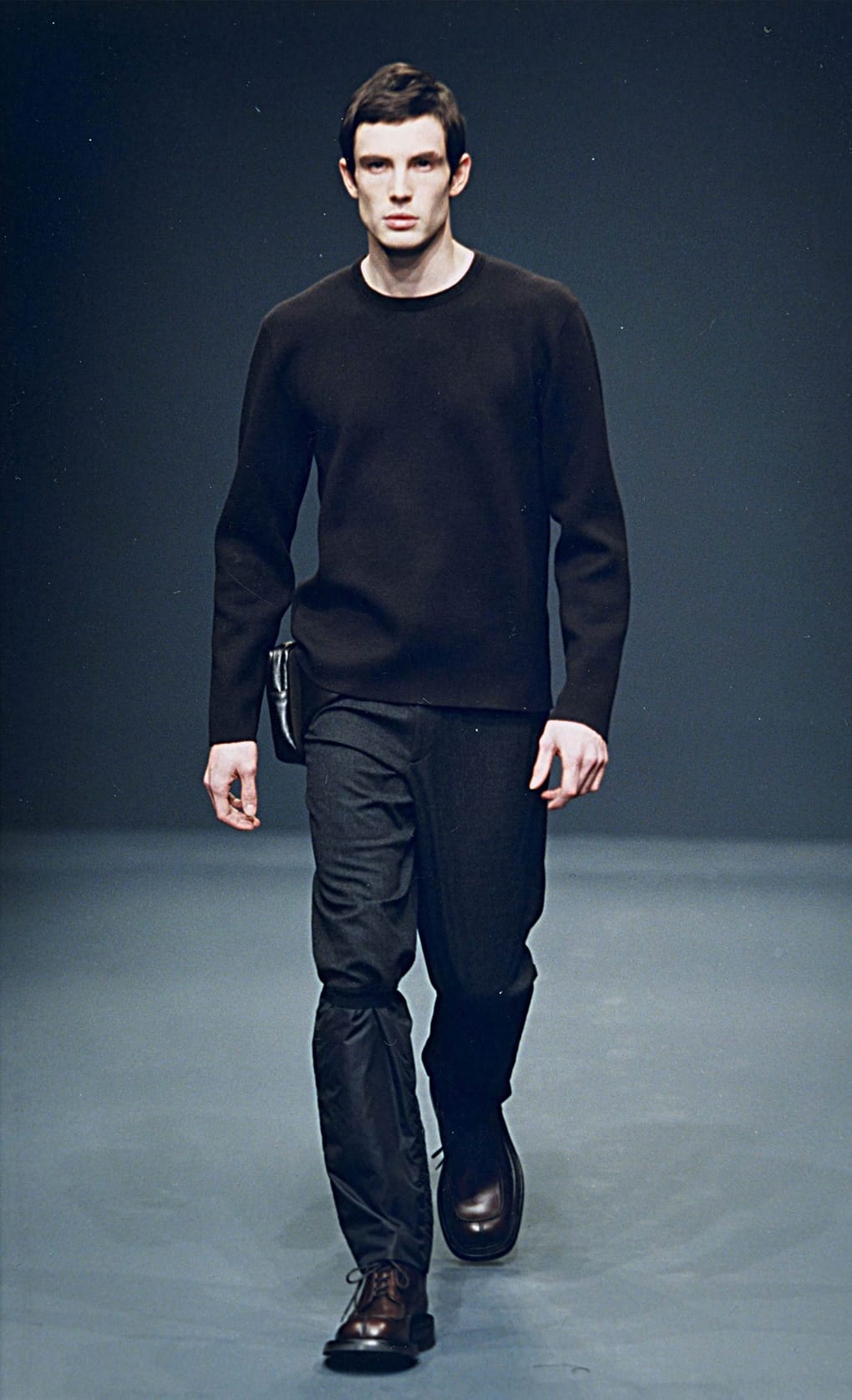 Prada Fall/Winter 1999 menswear runway look 33: black crewneck sweater and dark trousers with a small shoulder bag.