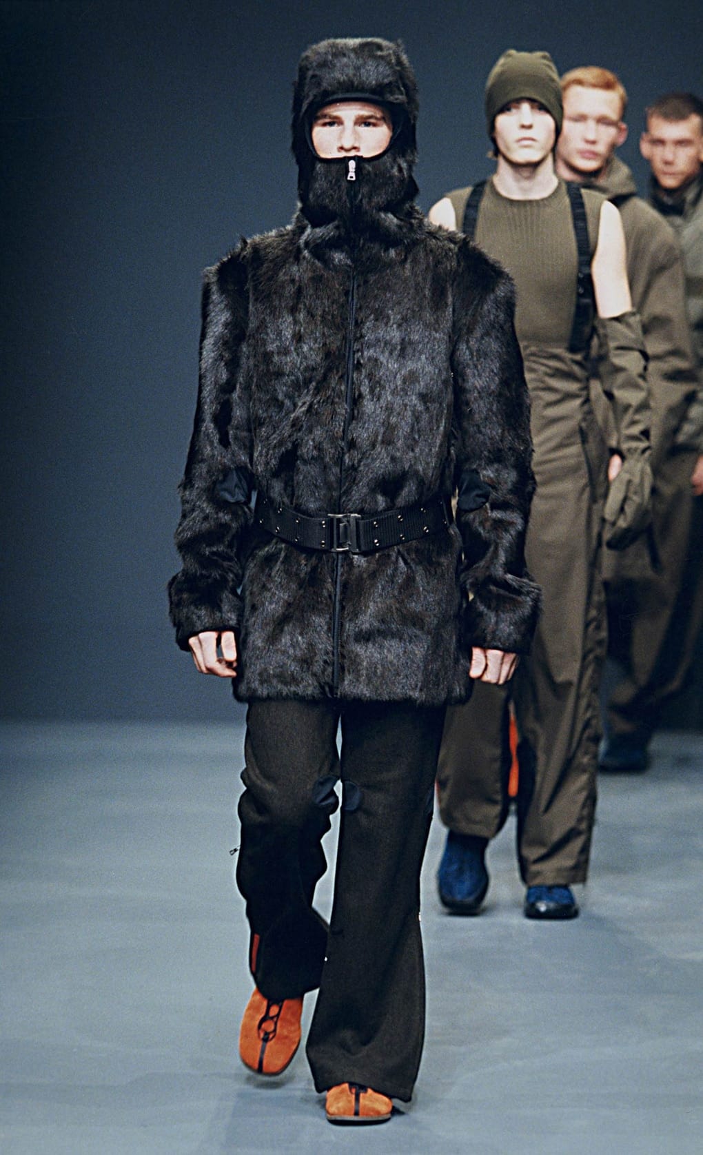 Prada Fall/Winter 1999 menswear runway look 36: black fur vest with hood, belt, dark trousers, and orange shoes.