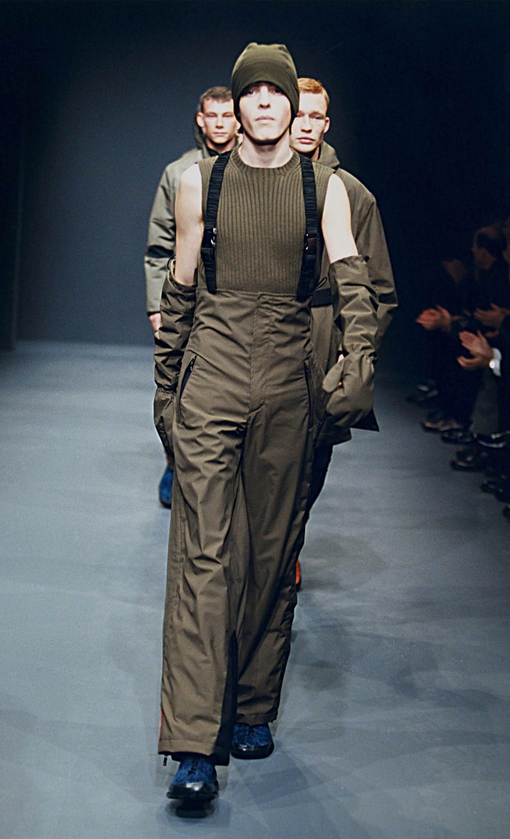 Prada Fall/Winter 1999 menswear runway look 37: olive technical layers with knit cap, harness straps, and dark trousers.
