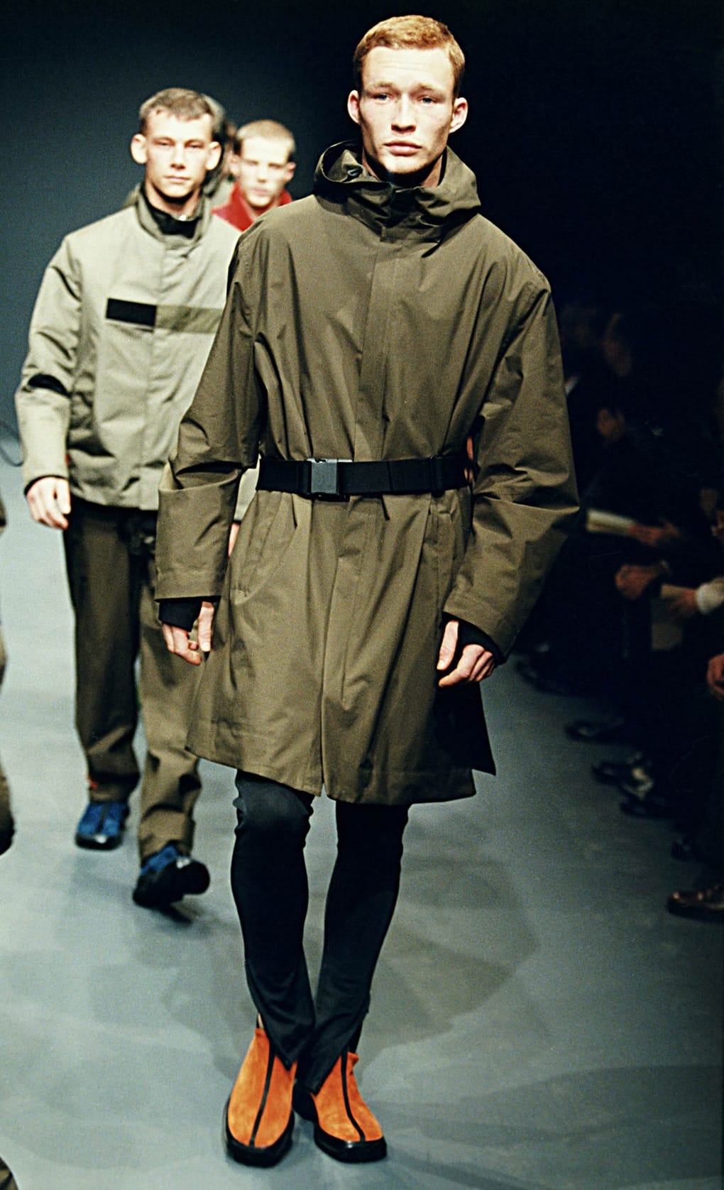 Prada Fall/Winter 1999 menswear runway look 38: olive belted hooded coat with black leggings and orange shoes.