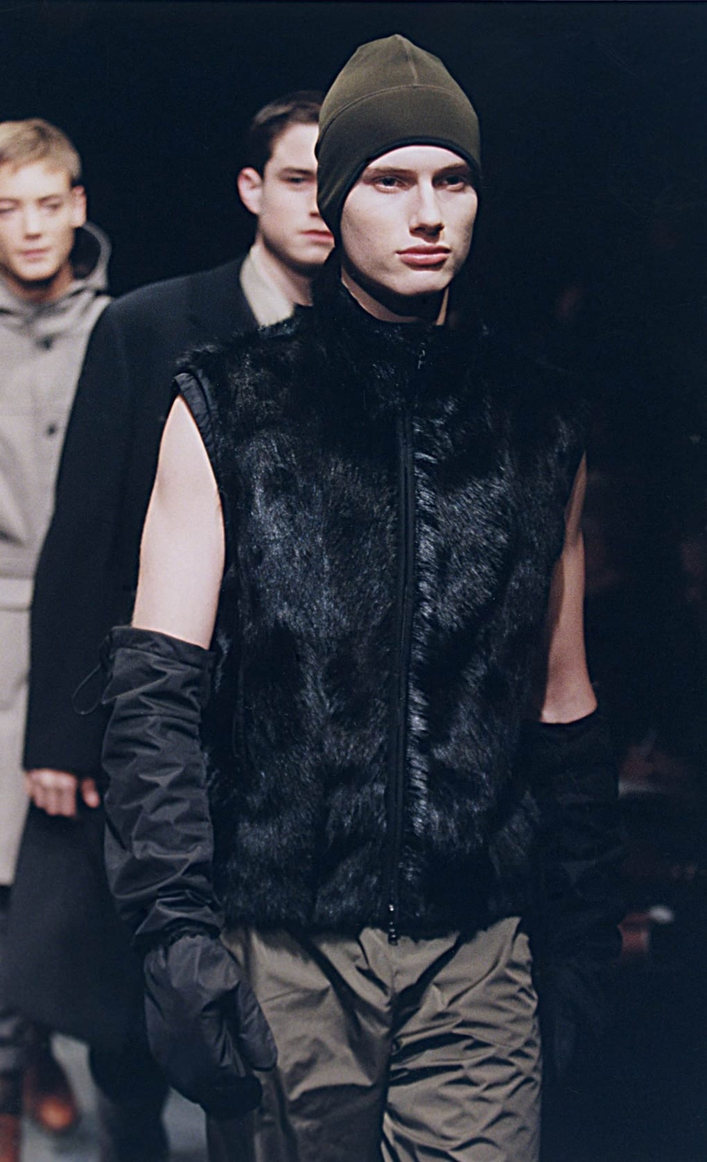 Prada Fall/Winter 1999 menswear runway look 55: black fur sleeveless top with detached sleeves and grey trousers.