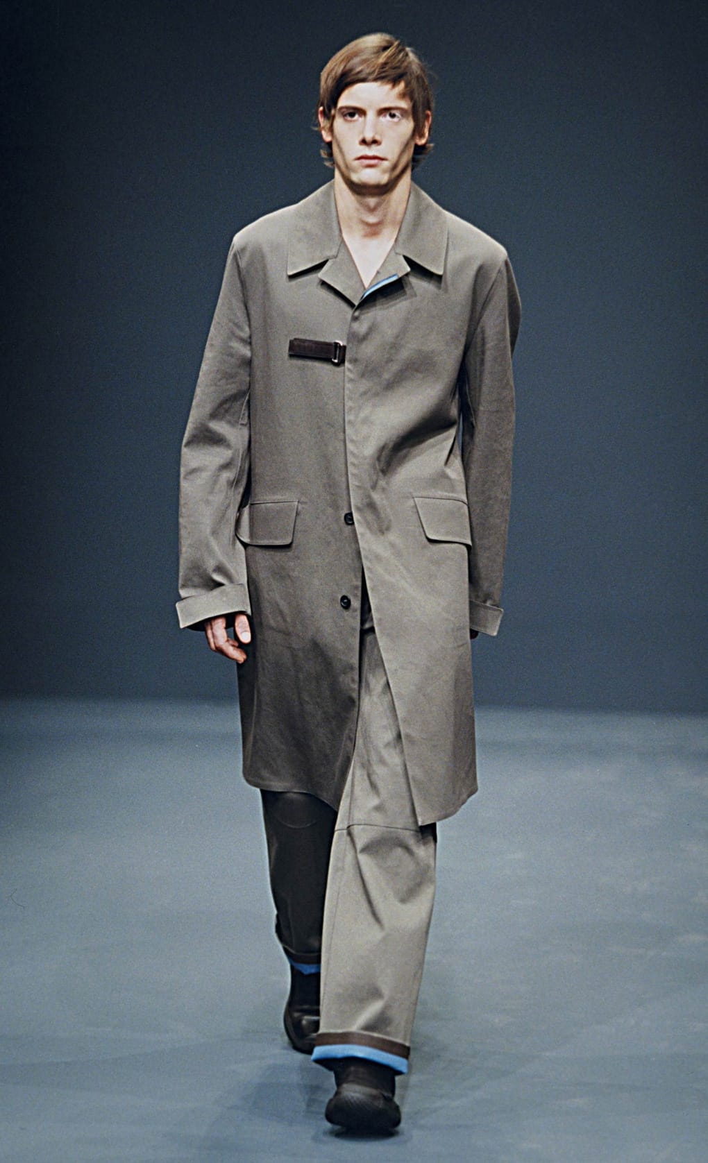 Prada Fall/Winter 1999 menswear runway look 60: grey long coat with matching trousers and layered blue cuffs.