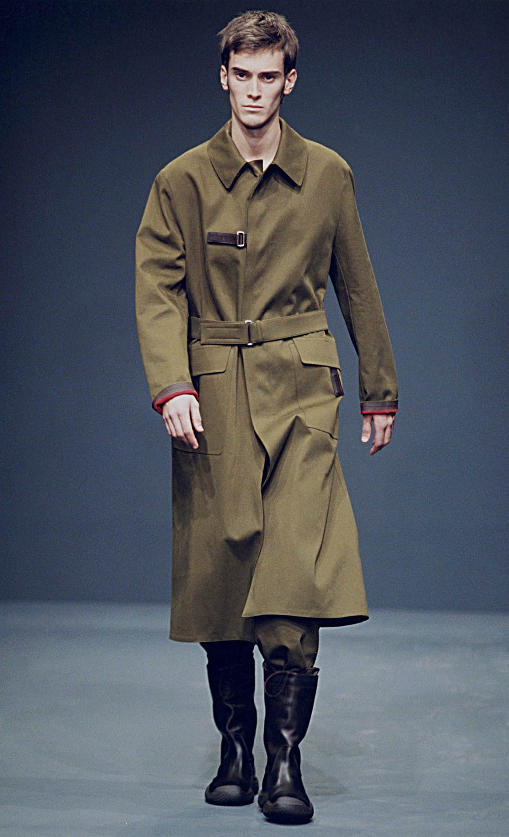 Prada Fall/Winter 1999 menswear runway look 67: olive belted long coat with dark cropped trousers and tall boots.