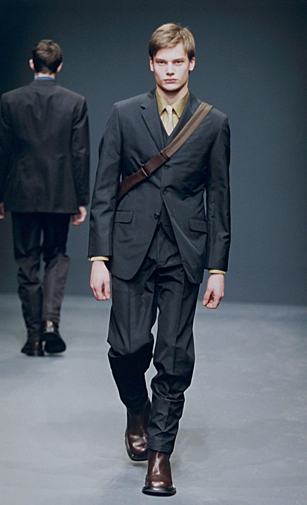 Prada Fall/Winter 1999 menswear runway look 72: dark suit with crossbody strap, olive shirt, and tall boots.