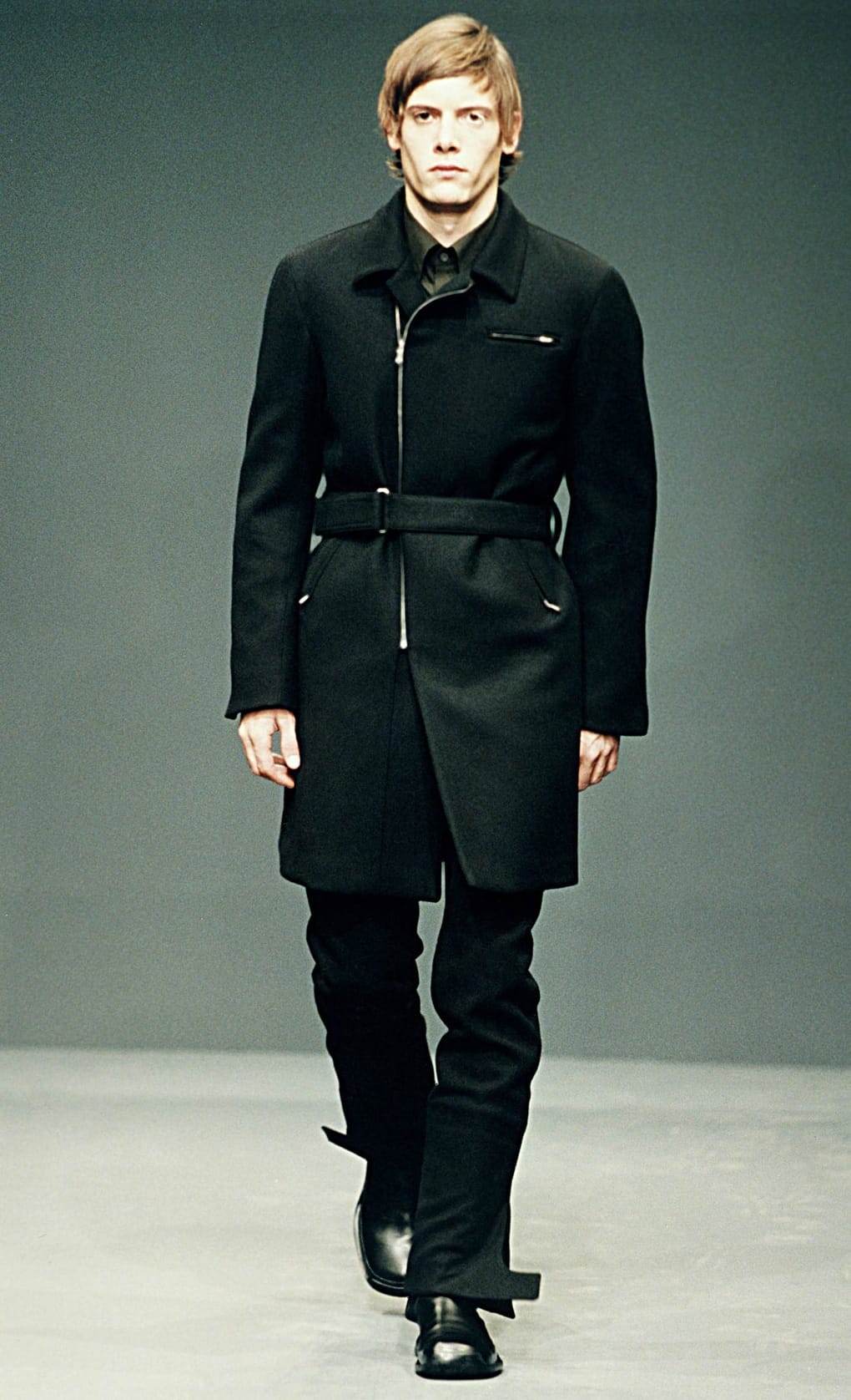 Prada Fall/Winter 1999 menswear runway look 77: belted black coat with dark trousers and tall boots.